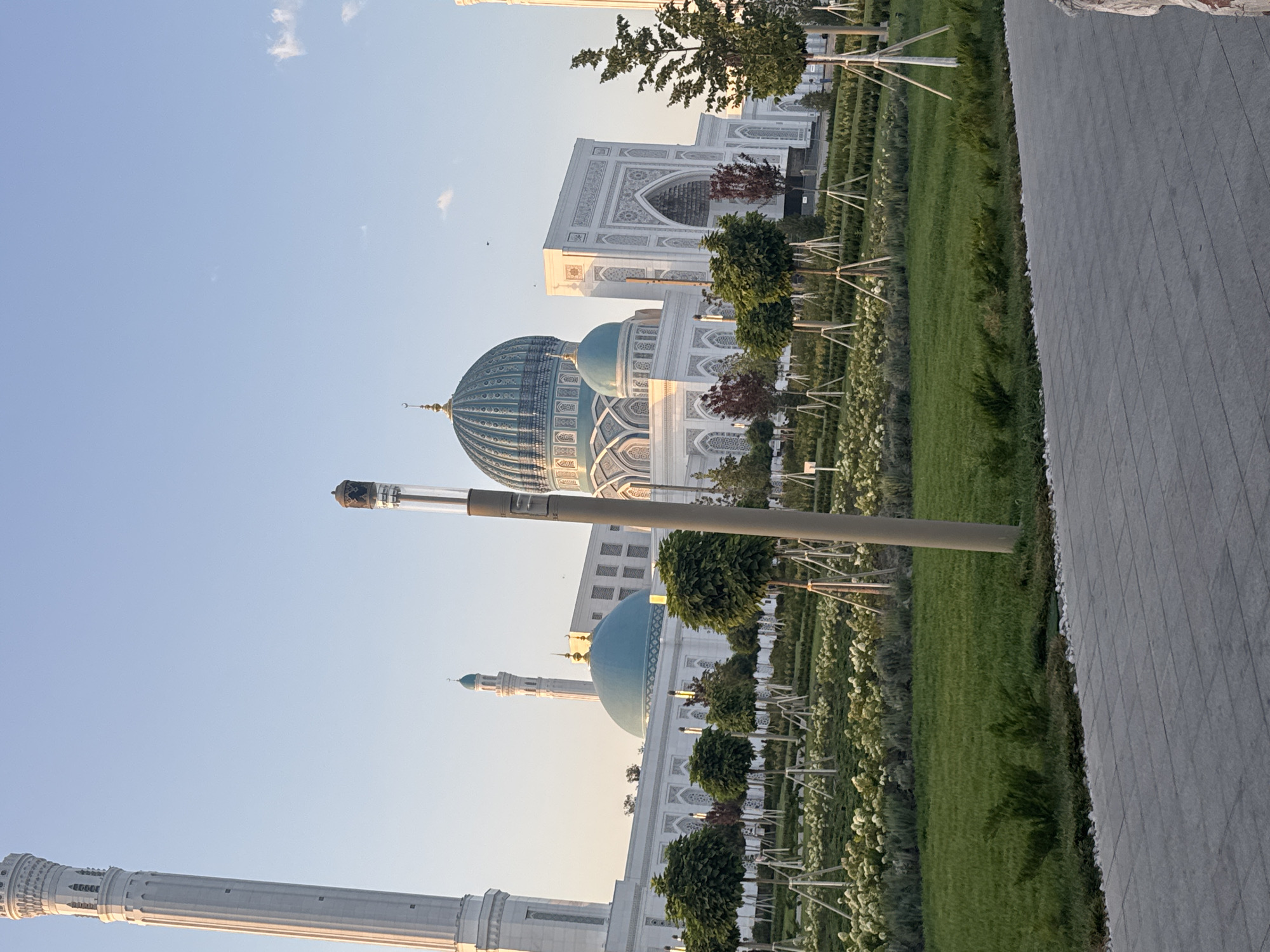 The image captures a view of the Hazrati Imam Complex in Tashkent, Uzbekistan, on a clear day. In the foreground, a paved walkway leads into a well-maintained garden with manicured grass, young trees supported by stakes, and beds of white flowers. A tall, cylindrical lamppost stands prominently in the garden. In the background, the distinctive white architecture of the Hazrati Imam Complex is visible, featuring multiple minarets, domes, and intricately decorated facades. One large, turquoise dome is particularly striking. The sky is a pale blue, with a few wispy clouds. The overall scene suggests a serene and well-preserved religious and cultural site.