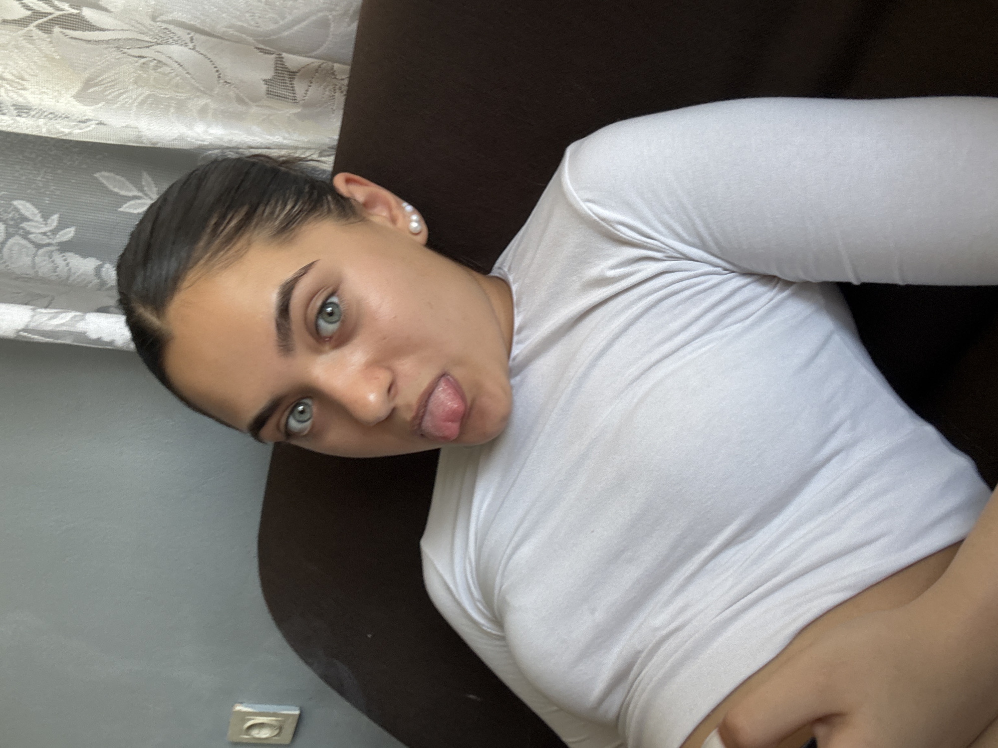 The image shows a close-up, chest-up view of a young woman with dark hair pulled back, blue-green eyes, and fair skin. She is wearing a white, long-sleeved shirt and has a pearl earring in her visible left ear. Her tongue is sticking out. She is positioned on a dark brown couch, with a grey wall and a lace curtain visible in the background. There is a light switch plate on the grey wall. The scene appears to be indoors, likely in a residential setting. There is no indication of the specific location within the city of Radievo, Bulgaria.