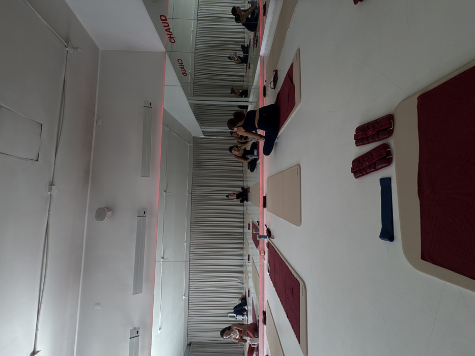 A fitness studio features mirrored walls and horizontal blinds. Rows of mats in shades of tan and burgundy are aligned on the floor. Women are seated on the mats, appearing to engage in exercises. Ankle weights and resistance bands are scattered on the floor near the mats. The wall is equipped with ventilation grills and the word "CHAUD" is written in uppercase letters on the mirror.
