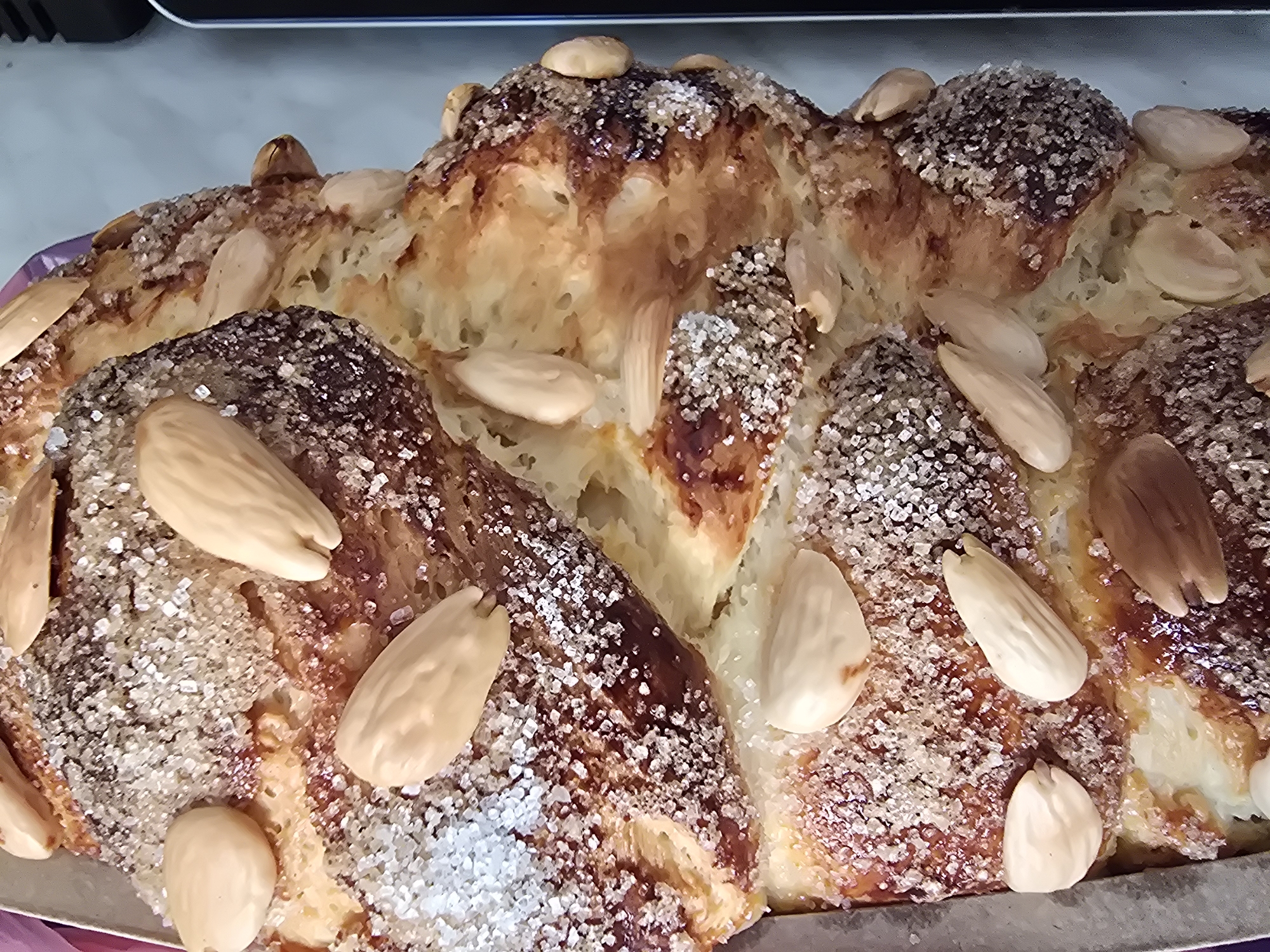 A close-up shot captures a sweet bread, possibly kozunak, adorned with whole almonds and coarse sugar crystals. The bread exhibits a golden-brown crust with lighter doughy interior sections visible where the bread has risen and expanded. The almonds are positioned artfully on top, while the sugar crystals create a sparkling effect across the crust's surface. No people are visible in the image. There are no indications in the photo that indicate it was taken in the city of Varna, Bulgaria.