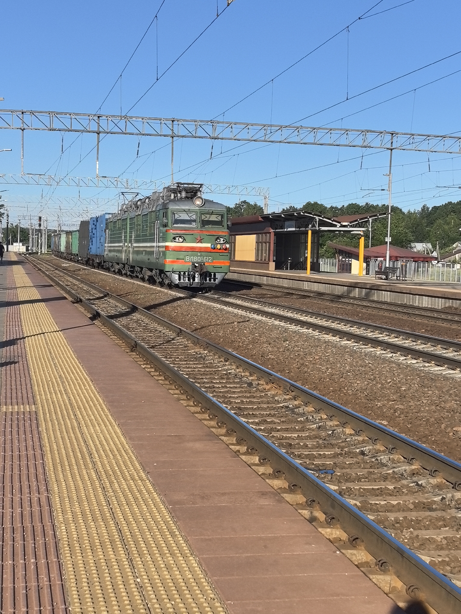 An exterior, eye-level, medium shot captures a green locomotive pulling a train through a railway station under a clear blue sky. The train is positioned on the right side of the frame, moving from left to right. The locomotive, with the number "ВЛ80-612" visible, is brightly lit by the sun. It is followed by several cargo containers in blue and green. The foreground is dominated by a train platform on the left, with a textured yellow strip running along its edge. Beside the platform are two sets of railway tracks. In the background, a small railway station building with a brown roof and yellow support columns is visible, along with trees and houses. The scene appears to be in Калодзішчанскі сельскі Савет, Belarus. A single person is walking away from the camera on the platform.