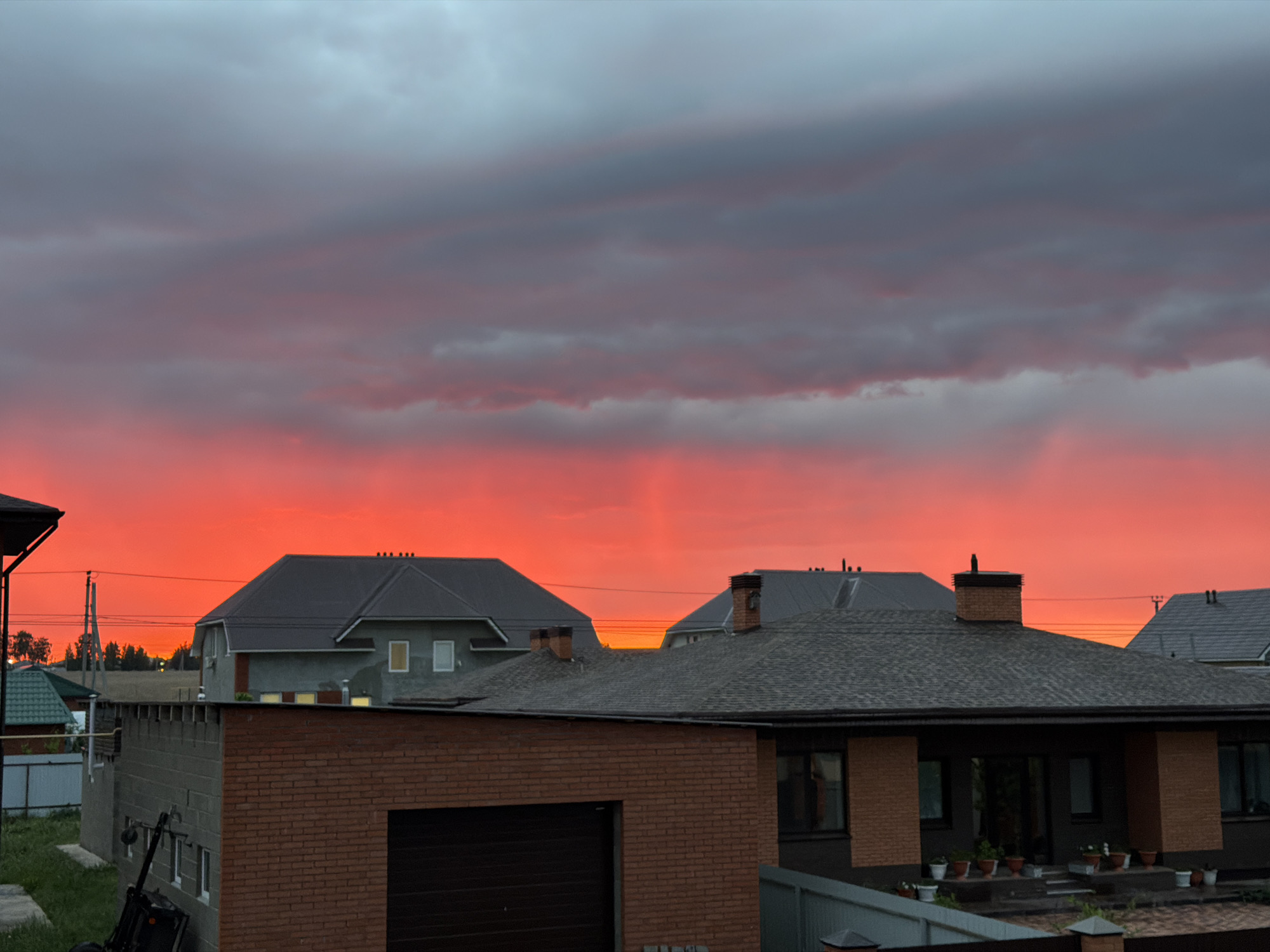The image shows a neighborhood in Большая Шильна, Russia, at sunset. The sky is a gradient of red and dark gray clouds. Several houses with gray roofs are visible, including one under construction with exposed brick. A brick garage is in the foreground. Utility poles are visible in the distance. There are potted plants near one of the houses.