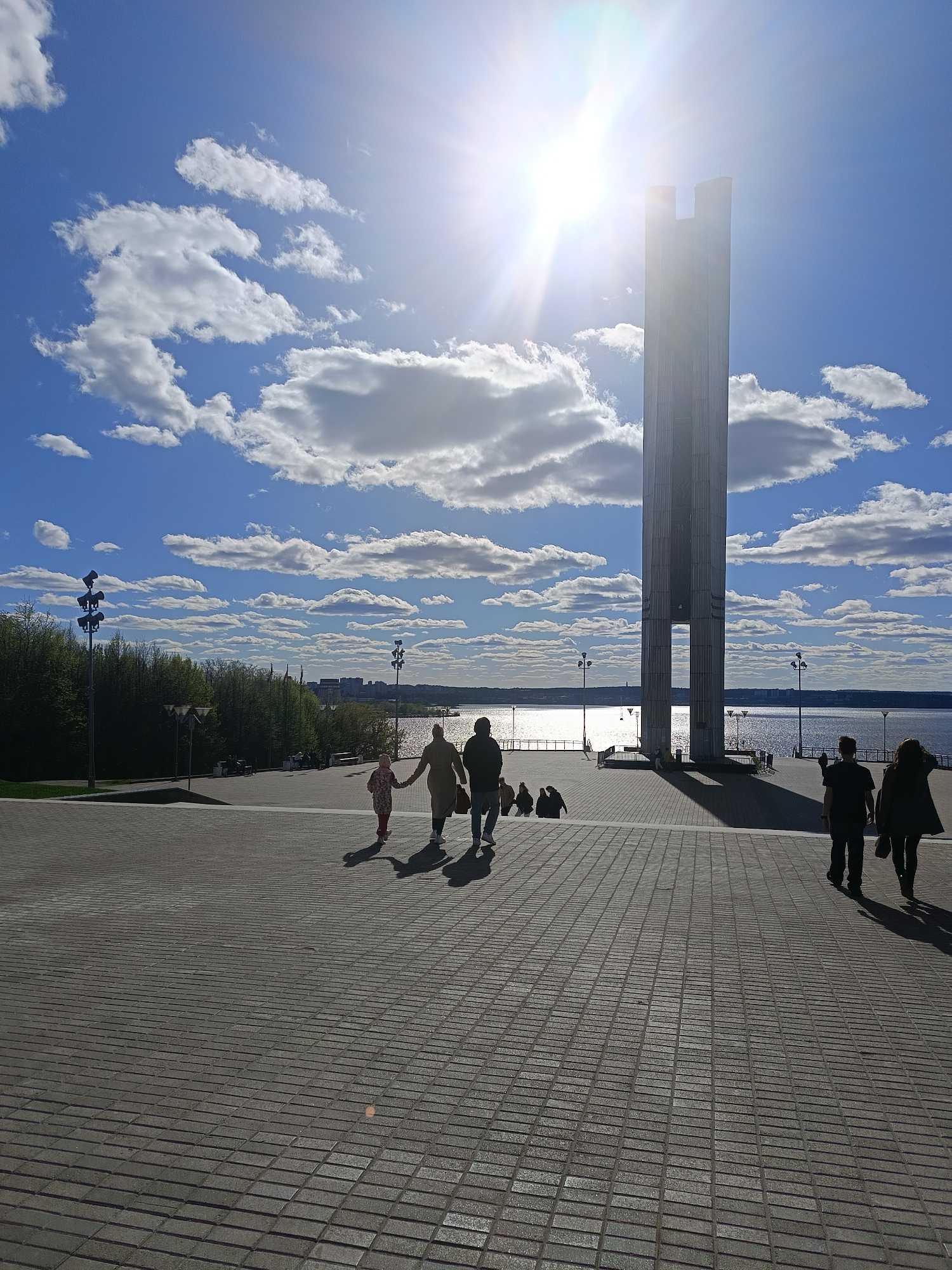 The image shows a wide-angle view of Victory Square in Izhevsk, Russia, under a bright, sunny sky with scattered clouds. A tall, rectangular monument stands prominently in the background, its structure reflecting the sunlight. Behind the monument is a body of water with a visible shoreline in the distance.
In the foreground, a paved area stretches out, leading towards the monument. Several people are walking on the pavement, some holding hands. Other people are sitting near the base of the monument. Trees and lamp posts are visible on the left side of the frame, alongside some buildings in the distance. The overall scene suggests a public gathering place or park area.