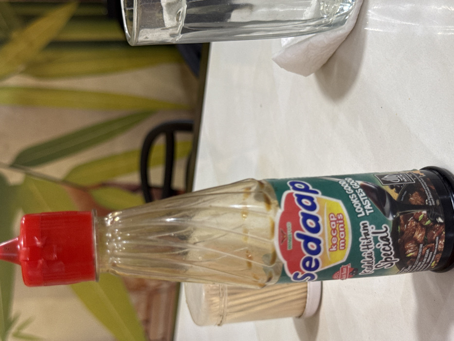 The scene, located in Payameuneng, Indonesia, depicts a close-up of a "Sedaap Kecap Manis Kedelai Hitam Special" sweet soy sauce bottle positioned on a white, reflective table surface. The clear plastic bottle features a ribbed upper section and a red dispenser cap. Its label is green, red, and yellow, displaying the brand name, product description, a graphic of a dark, sauced dish with black soybeans, and the phrase "LOOKS GOOD TASTES GOOD," along with a Halal certification logo. To the left of the bottle, a translucent cylindrical container filled with wooden toothpicks is visible. To the right, a clear, ribbed drinking glass and a crumpled white napkin rest on the table. The background is out of focus, revealing blurry green and yellow vertical patterns, consistent with decorative bamboo imagery, and a dark, curved object resembling a chair back.