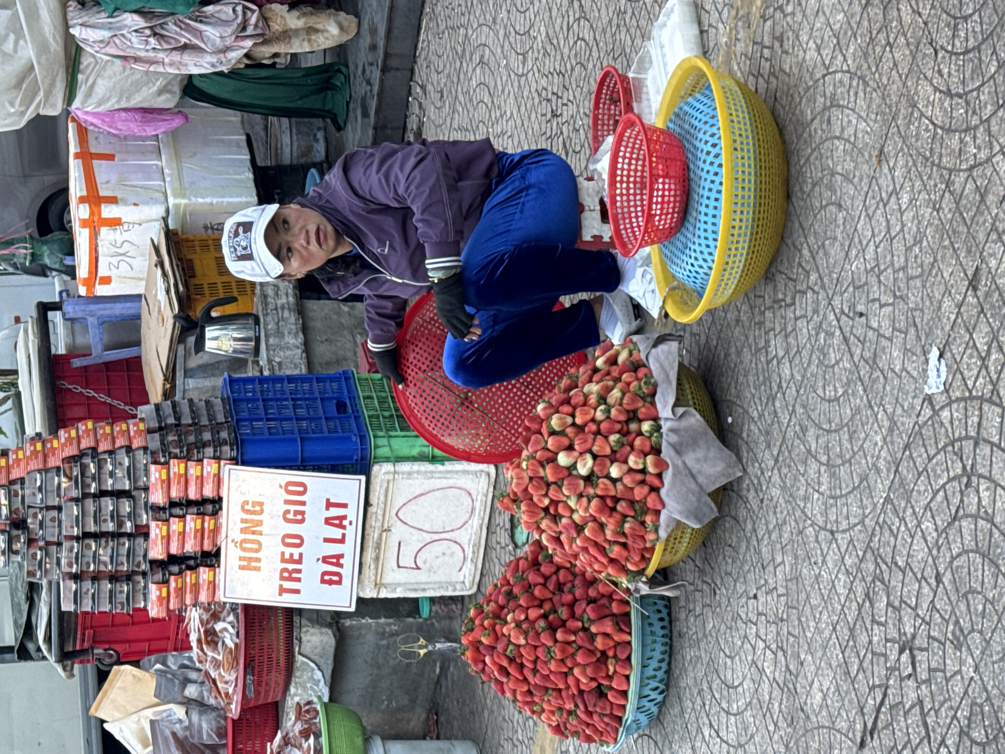 In Da Lat, Vietnam, a woman wearing a purple jacket, blue pants, a white baseball cap, and black gloves is seated on a small red stool. She is positioned in front of a stall displaying large quantities of strawberries. Two prominent baskets filled with strawberries are in the foreground. Behind her, a white sign with Vietnamese text reading "HONG TREO GIO DA LAT" and the number "50" is visible. Various crates and containers are stacked in the background, indicating a market setting. The ground is paved with patterned tiles.