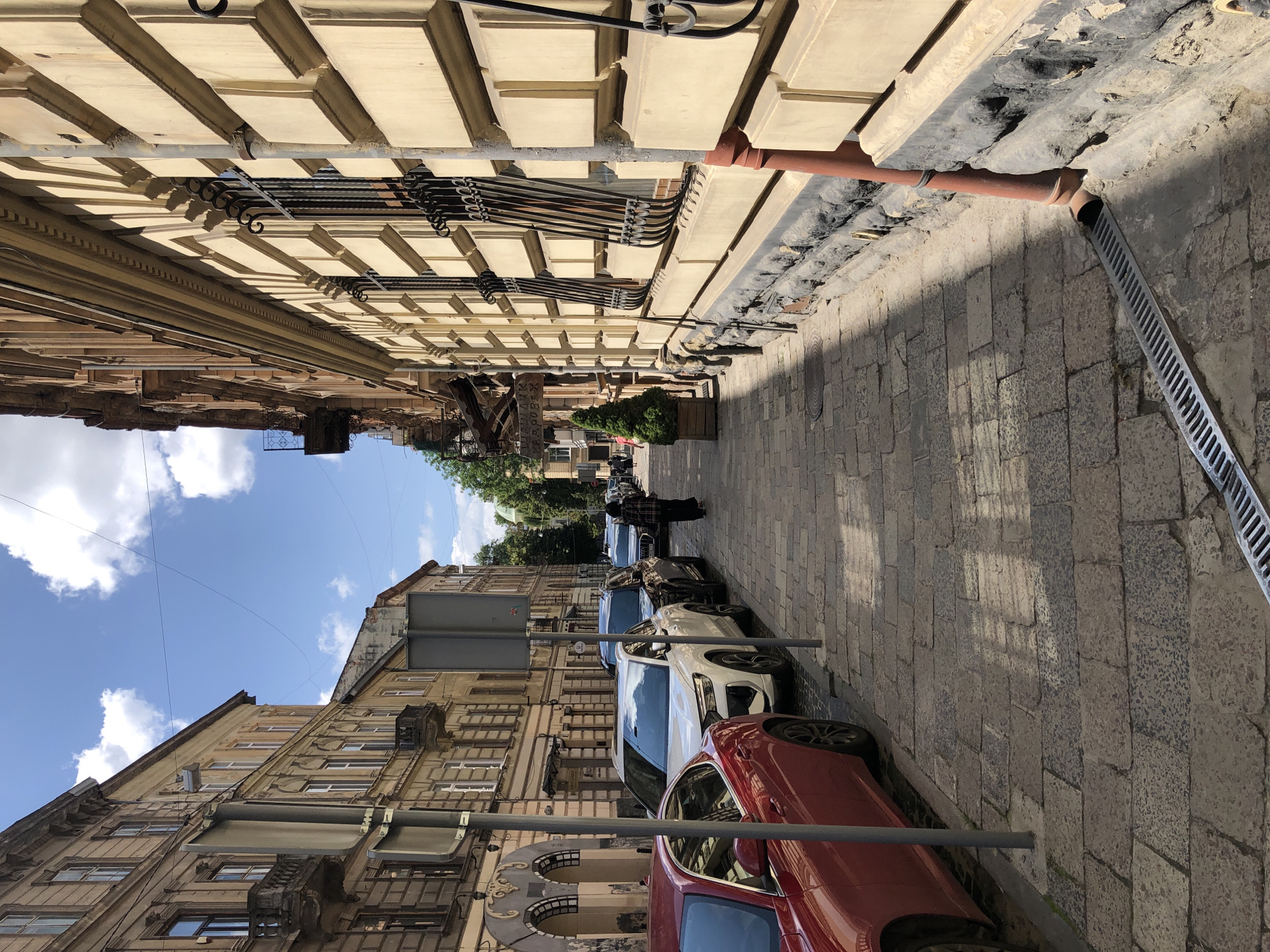 The image depicts a narrow street in the historic city center of Lviv, Ukraine. The perspective is from the street level, looking down the cobblestone pavement. On the right, a large building with ornate yellow stucco facade and wrought-iron window grates lines the street. A rusty-colored drainpipe runs vertically down the side of this building, disappearing into a drainage grate on the sidewalk. The building on the left is also multi-story, with a more muted beige facade, visible balconies, and arched doorways at street level.
Several cars are parked along the left side of the street. A bright red car is in the foreground, followed by a white Audi SUV, a black SUV, and other vehicles further down the street. A person in dark clothing, possibly a woman with a patterned scarf, walks away from the camera further down the street on the right side. The sky is blue with scattered white clouds, indicating a sunny day. The overall impression is one of a well-preserved, classic European city street scene.