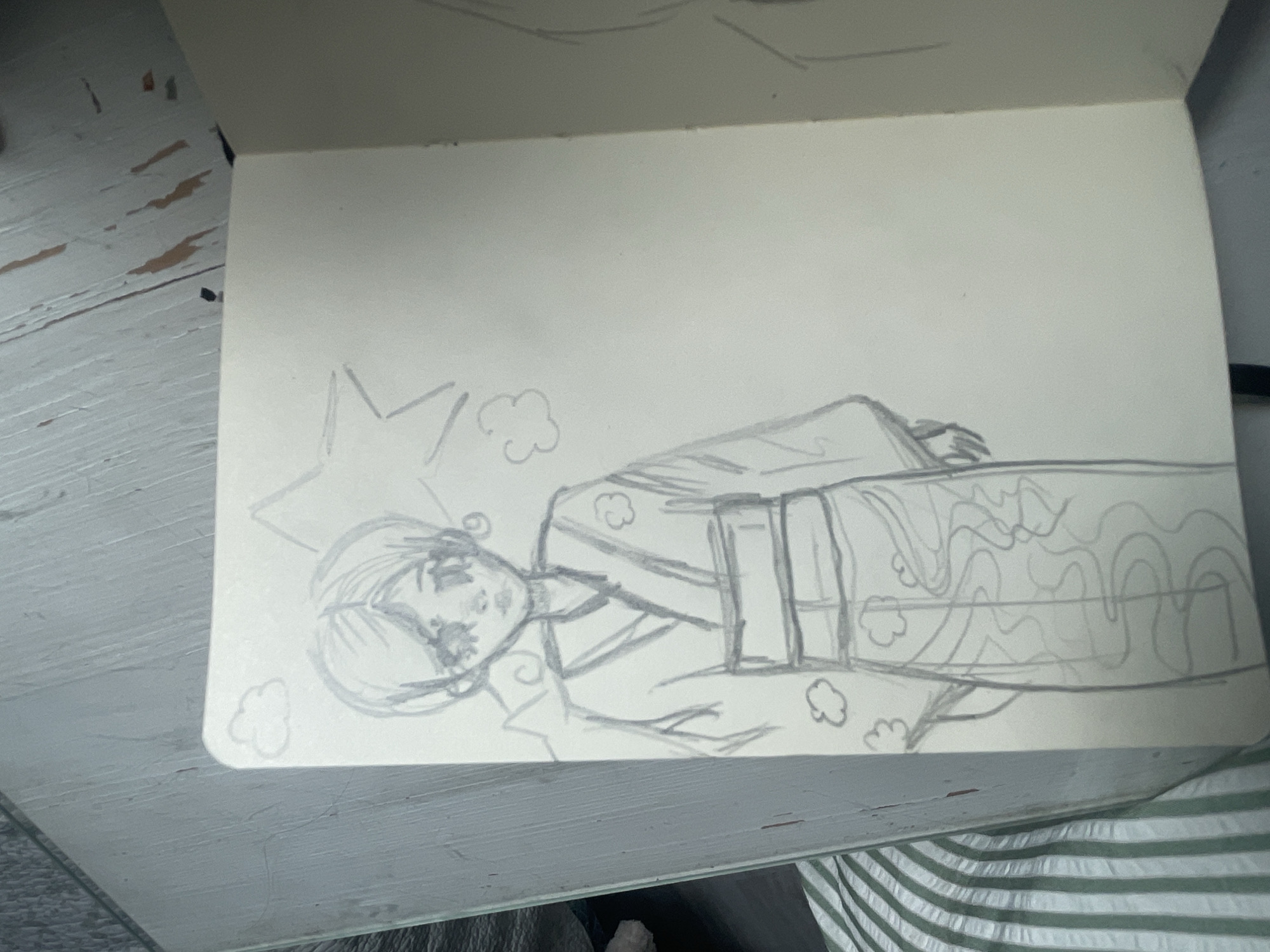 The image shows a sketch of a person in a kimono, drawn in a notebook. The sketch is in black and white and appears to be in a simplified, anime-inspired style. The person has large eyes and short hair. The kimono has a pattern of clouds and waves. Behind the person, there is a large star shape and some smaller cloud shapes. The notebook is open, and the page to the right of the sketched page shows another unfinished sketch. The notebook is placed on a surface that appears to be made of wood, with a striped fabric peeking out from underneath. There are no discernible elements in the image that would indicate the location as being in the city of Мухавецкі сельскі Савет, Belarus.