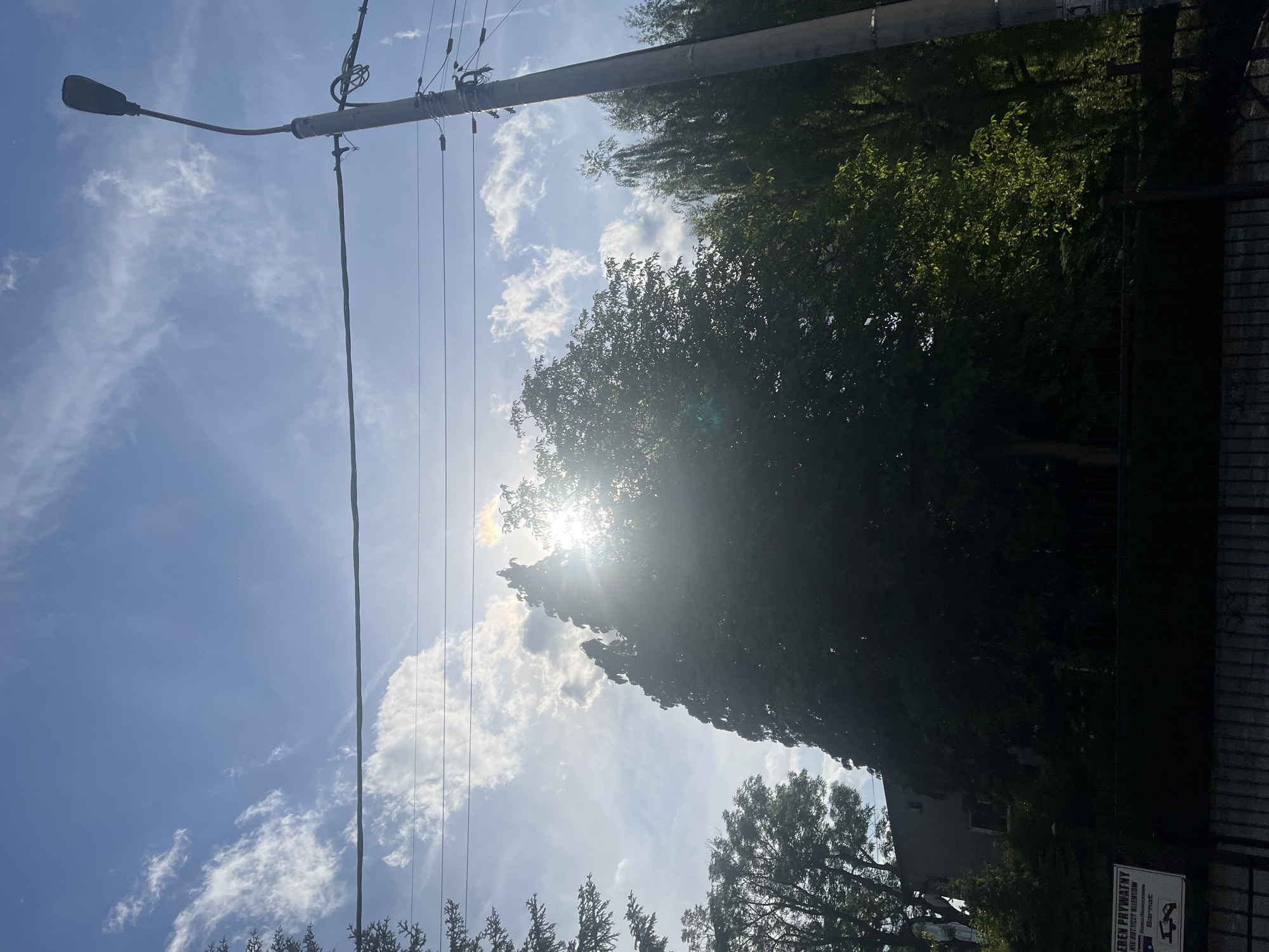 The image shows a daytime scene in Unknown, Unknown. A utility pole with a streetlight and power lines extends across the upper left of the frame. The sky is visible with patches of clouds. Trees with dense foliage are present in the upper right and center of the image, with the sun positioned behind them. A building is partially visible behind the trees near the lower right, and a sign written in Polish is attached to a fence.