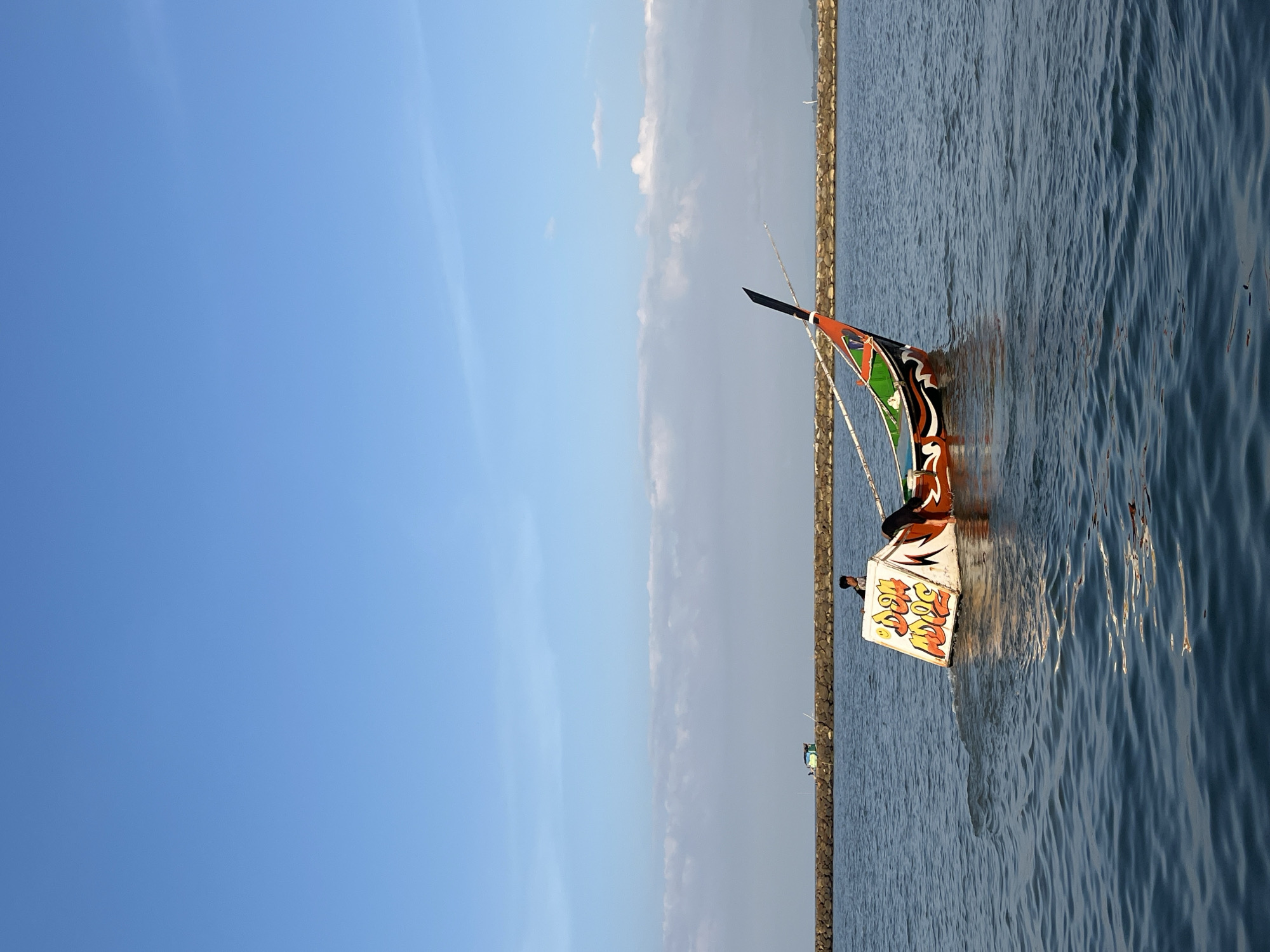 A traditional fishing boat with vibrant orange, green, and black decorations is positioned on the water. A person is standing on the boat, facing towards the back. The boat has a white panel with orange and yellow lettering on its side. The boat is moving, creating a wake behind it. In the background, a stone breakwater extends across the water, with a small green structure visible on top. The sky is a clear blue with thin white clouds. The scene appears to be a coastal area in Meunasah Jurong, Indonesia.