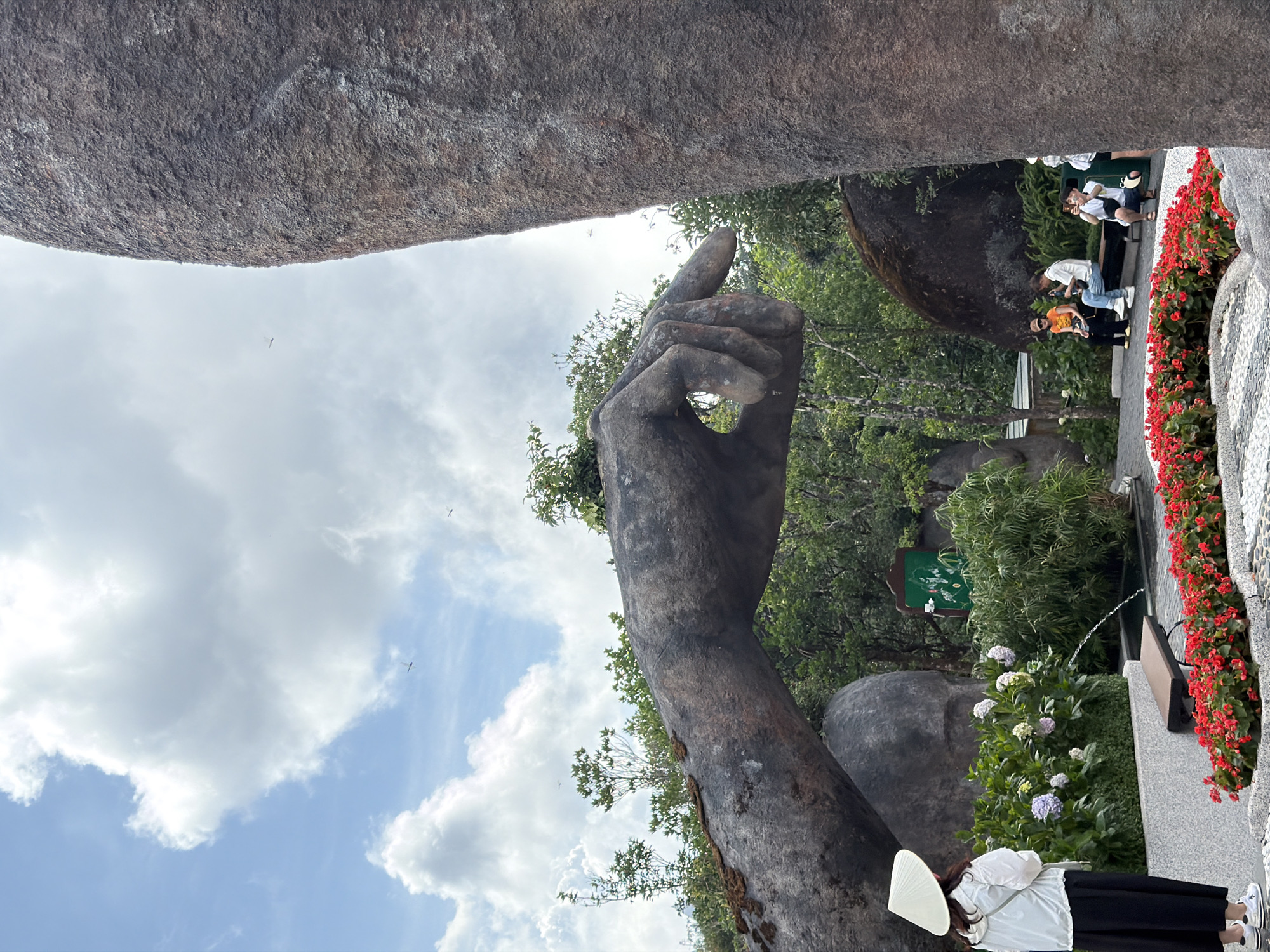 This is a vertical outdoor shot taken at the Golden Bridge in Đà Nẵng, Vietnam. The image captures a section of the massive stone hand sculpture, with a large rock formation above it. The sky is visible between the rock and the pointing finger of the hand. A group of people can be seen in the background along a paved pathway lined with red flowers. There is also a woman in a conical hat with her back turned. Hydrangeas and other greenery are also visible behind the hand.