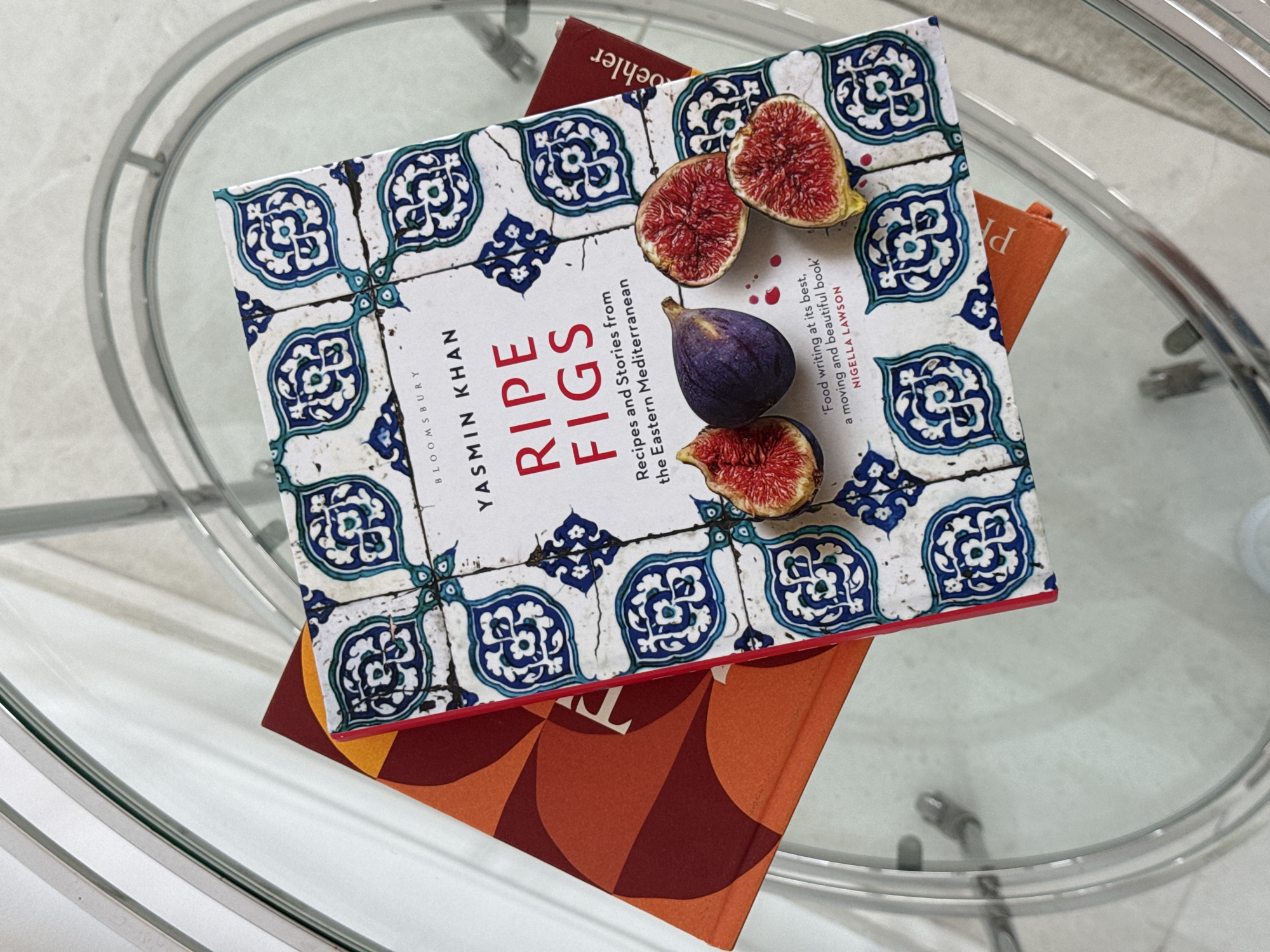 The image depicts a stack of books on a glass-topped table. The uppermost book, titled "Ripe Figs" by Yasmin Khan, features an image of figs on its cover. Underneath this book, another book can be partially seen, with a maroon and orange color scheme. The surrounding environment appears to be an indoor setting, given the nature of the furniture. Based on the location specified, the scene is likely within the city of Ghadeer Al Tayr, United Arab Emirates, but nothing in the image confirms this.