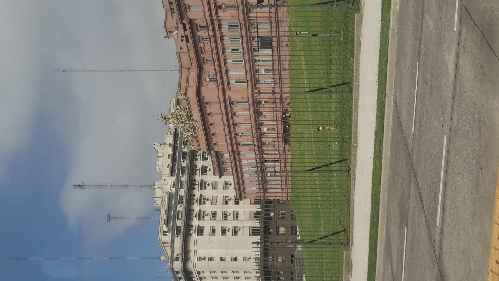 The image depicts the Casa Rosada, the executive mansion and office of the President of Argentina, located in Buenos Aires. Adjacent to the building is a green space, separated from the street by a black iron fence. The street has a yellow curb and white lane markings. In the background is a blue sky with clouds. Communication towers are seen behind the Casa Rosada.