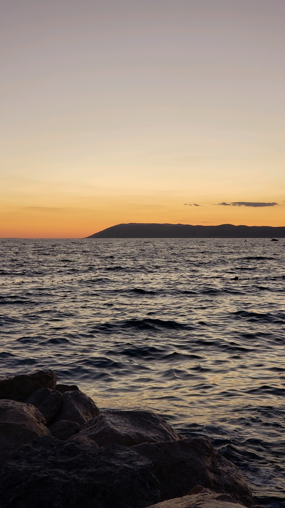 The image depicts a serene seascape at sunset in Općina Tučepi, Croatia. The foreground is dominated by a cluster of large, dark, rough-textured rocks forming a natural barrier along the shoreline. Beyond the rocks, the dark blue water of the Adriatic Sea stretches towards the horizon, its surface rippled by gentle waves reflecting the warm hues of the sky. In the mid-ground, a thin line of buoys or markers is visible on the water's surface. In the distance, a silhouetted mountain range forms the backdrop, its dark mass contrasting with the soft gradient of the sky. The sky itself transitions from a pale, almost lavender grey at the top to a vibrant orange and yellow near the horizon, indicative of a setting sun. A single, small, dark cloud is visible in the upper right portion of the sky, and a faint outline of a boat is present on the far right horizon. The overall impression is one of tranquility and natural beauty during twilight.