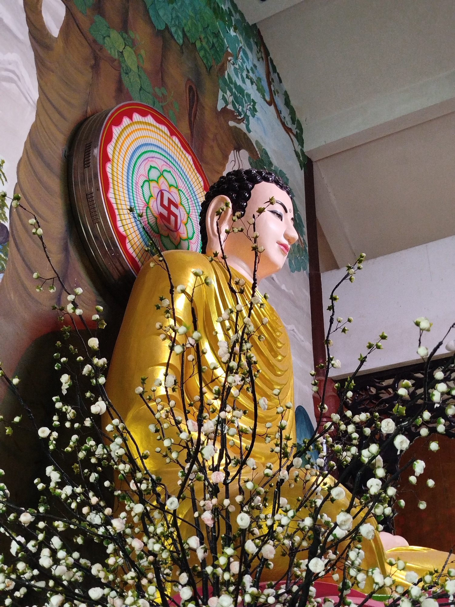 Captured indoors, the image showcases a statue of Buddha in a temple, likely located in Xuân Trường - Đà Lạt, Vietnam. The golden-hued Buddha sits serenely, positioned in front of a wall that is decorated with an illustration of a tree. A circular, decorative element with a swastika symbol is placed behind the statue. In the foreground, a branch laden with small white flowers partially obscures the view.