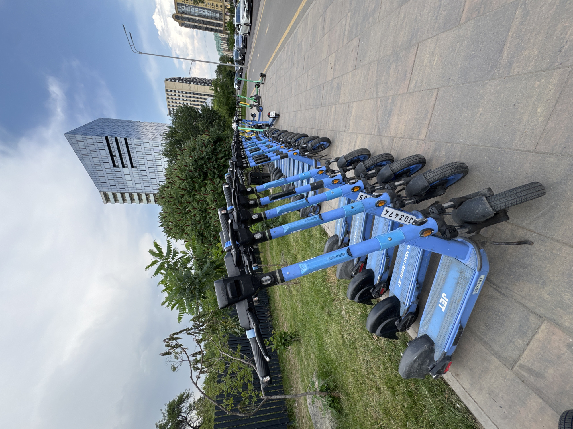 A row of electric scooters are parked on a sidewalk next to a grassy area with trees and a black fence. In the background, there are modern buildings, a street with cars, and a lamp post. The sky is partly cloudy. The scooters are blue with "JET" branding visible on their decks. One scooter has a license plate that reads "J033474". Another scooter has the number "257-933" printed on its deck. The overall scene suggests an urban environment in Almaty, Kazakhstan.