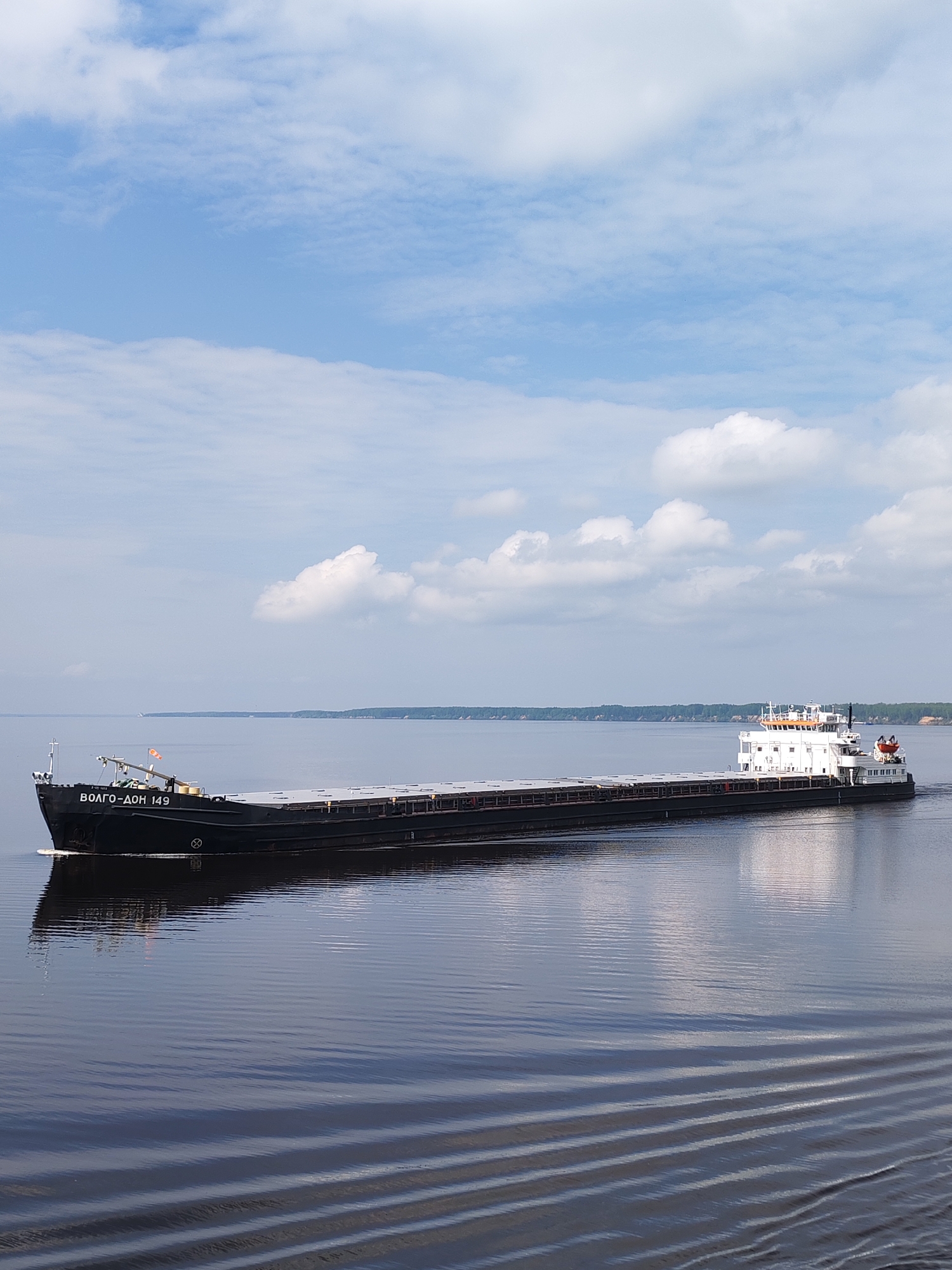 The image shows a large cargo ship, identified as "ВОЛГО-ДОН 149" (VOLGO-DON 149), sailing on a body of water, likely the Volga-Don Canal. The ship is dark in color with a white superstructure at the stern. It appears to be moving, as evidenced by the wake visible in the water. The sky is partly cloudy with a mix of blue sky and white clouds. In the distance, a shoreline with trees is visible. The water is relatively calm, with slight ripples reflecting the sky. There are no people visible in the frame. The location is likely near Gladyshevo, Russia, based on the ship's name suggesting a connection to the Volga-Don Canal.