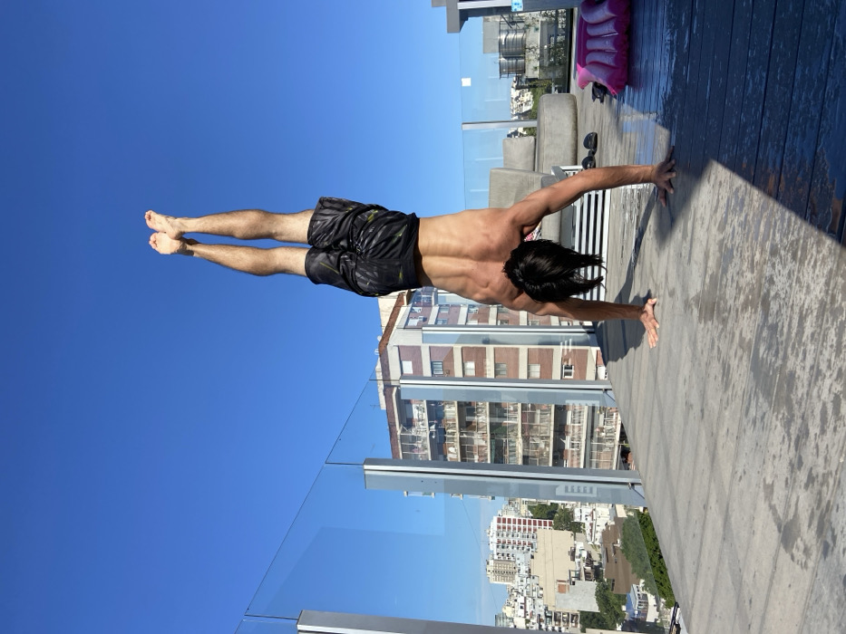 A shirtless man in swim trunks does a handstand on the edge of a rooftop. The blue sky is behind his legs while buildings and a city landscape are visible behind him. A glass railing protects the edge of the rooftop. A wooden deck is to the right, with furniture further back on the rooftop. The scene is shot in bright daylight.