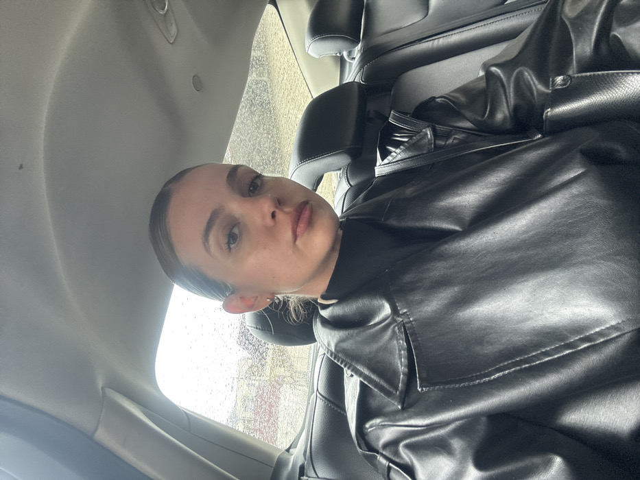 The image depicts a female subject seated in the rear passenger compartment of a vehicle in an unknown city, Unknown. The subject, a woman appearing to be in her mid-20s to early 30s, has light brown hair pulled back and a neutral facial expression while looking directly at the camera. She is wearing a black turtleneck top and a black faux-leather trench coat. A small hoop earring is visible in her left ear. The vehicle's interior features black leather seats with headrests and a grey fabric headliner. Through the rear window, precipitation is visible on the glass, indicating rain or wet weather conditions. The external background is indistinct due to blur and water on the window, showing vague shapes consistent with distant urban or suburban structures.