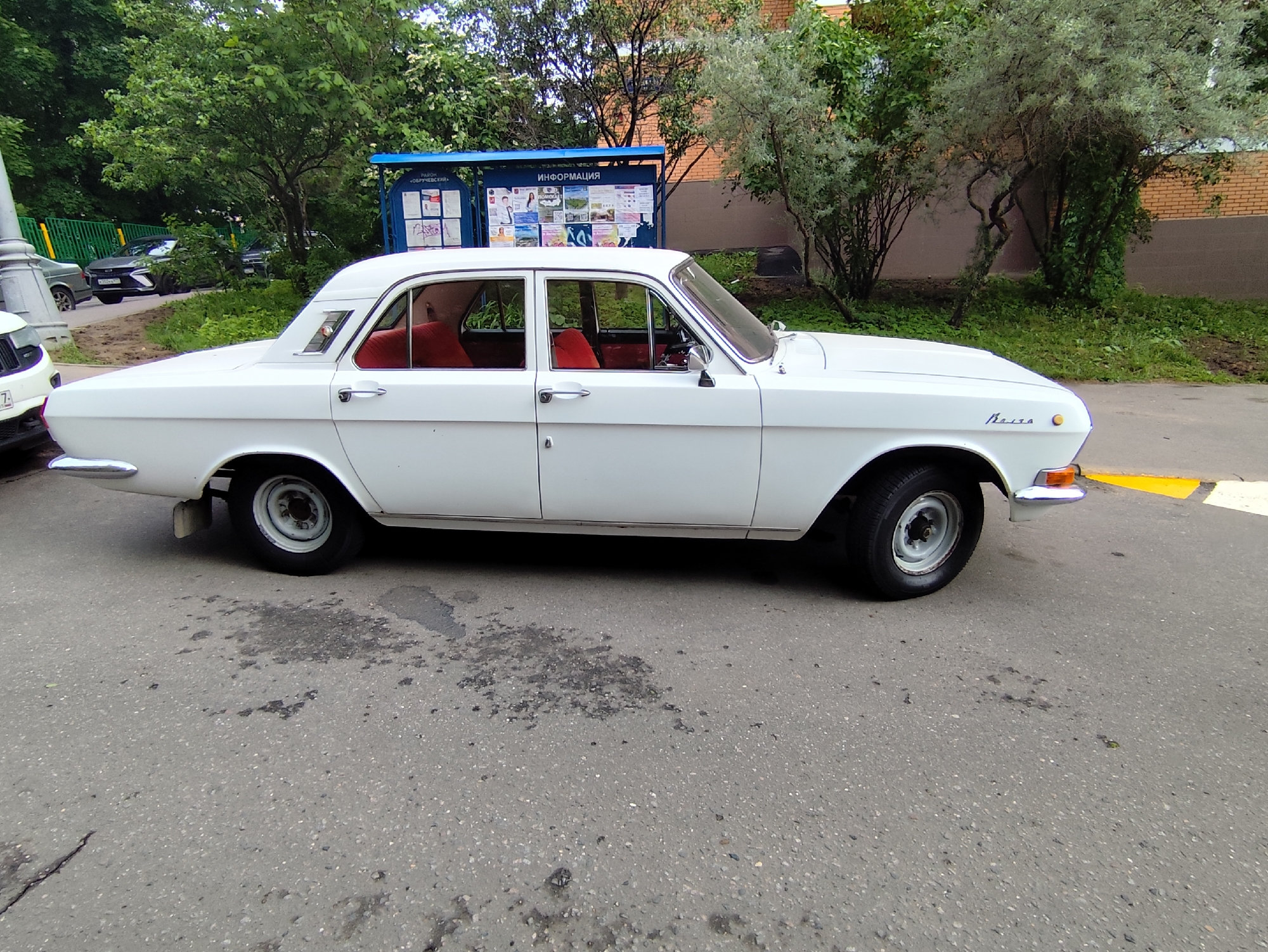 A white GAZ-24 Volga sedan is parked on an asphalt street, likely in Moscow, Russia. The car has red interior and gray wheels. Behind the car, there is a blue public information kiosk with the word "ИНФОРМАЦИЯ" (Information) visible on the top panel. To the left, partially obscured by trees, there are other parked vehicles. The background also includes trees, a brick building, and green bushes. The asphalt street is patchy with visible dark stains and repairs.