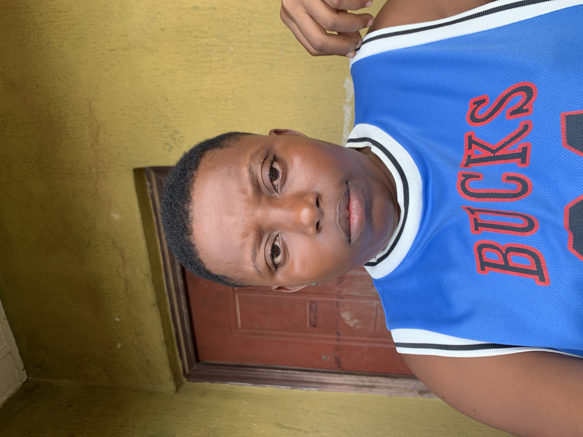 A medium shot depicts a person wearing a blue basketball jersey with "BUCKS" printed on it. The person is positioned in front of a brown door set in a yellow-painted wall. The location is Ikorodu, Nigeria.