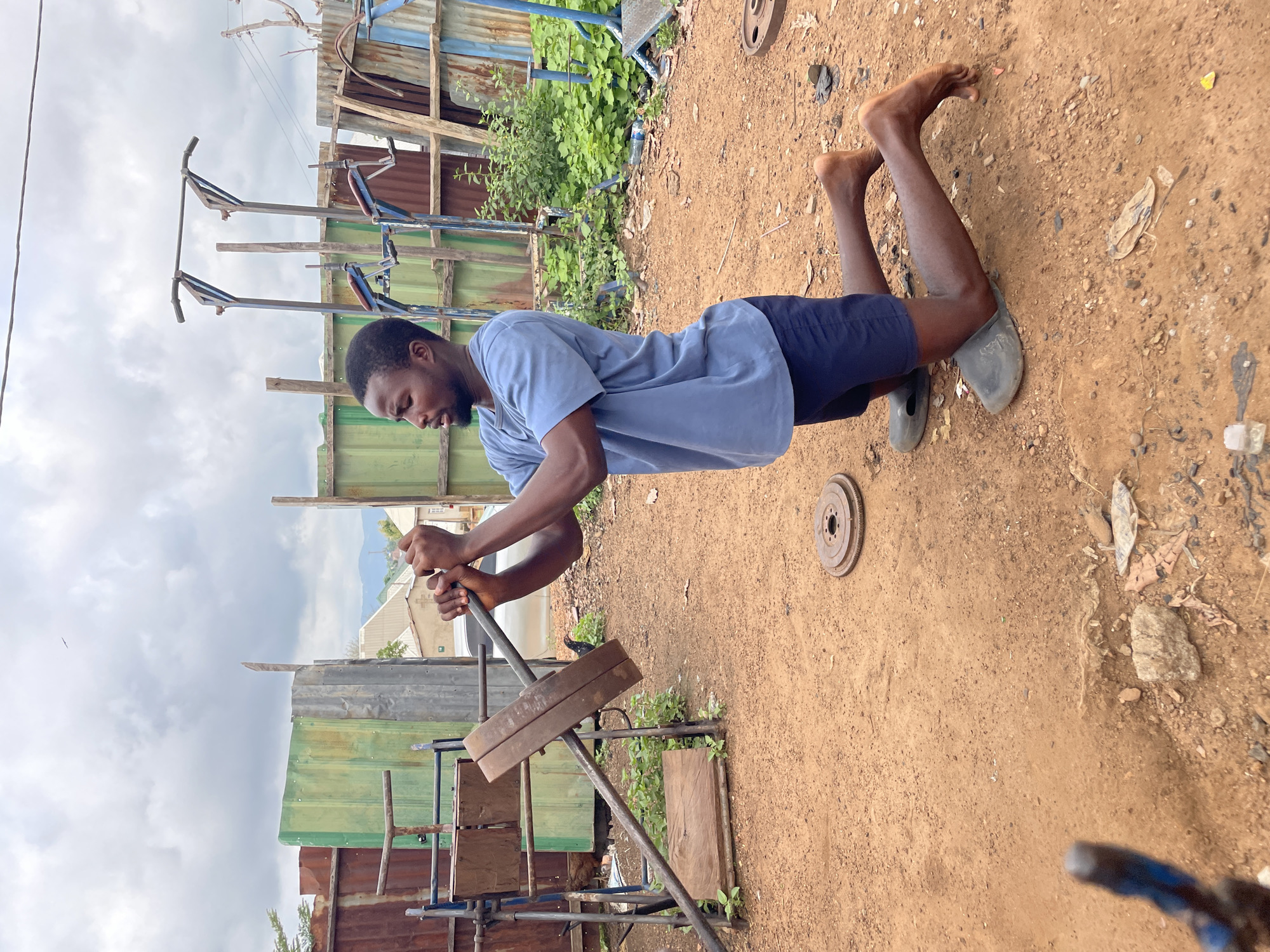 A man exercises in an outdoor gym in Akwanga, Nigeria. The man is light-skinned African and wears a blue t-shirt, blue shorts, and sandals. He is bent over, grasping a bar on a homemade weight-lifting machine with metal weights. The ground is dirt. Scrap metal and wood form the gym structure, with green and blue corrugated metal fencing in the background. In the distance, houses and trees are visible. The sky is overcast.
