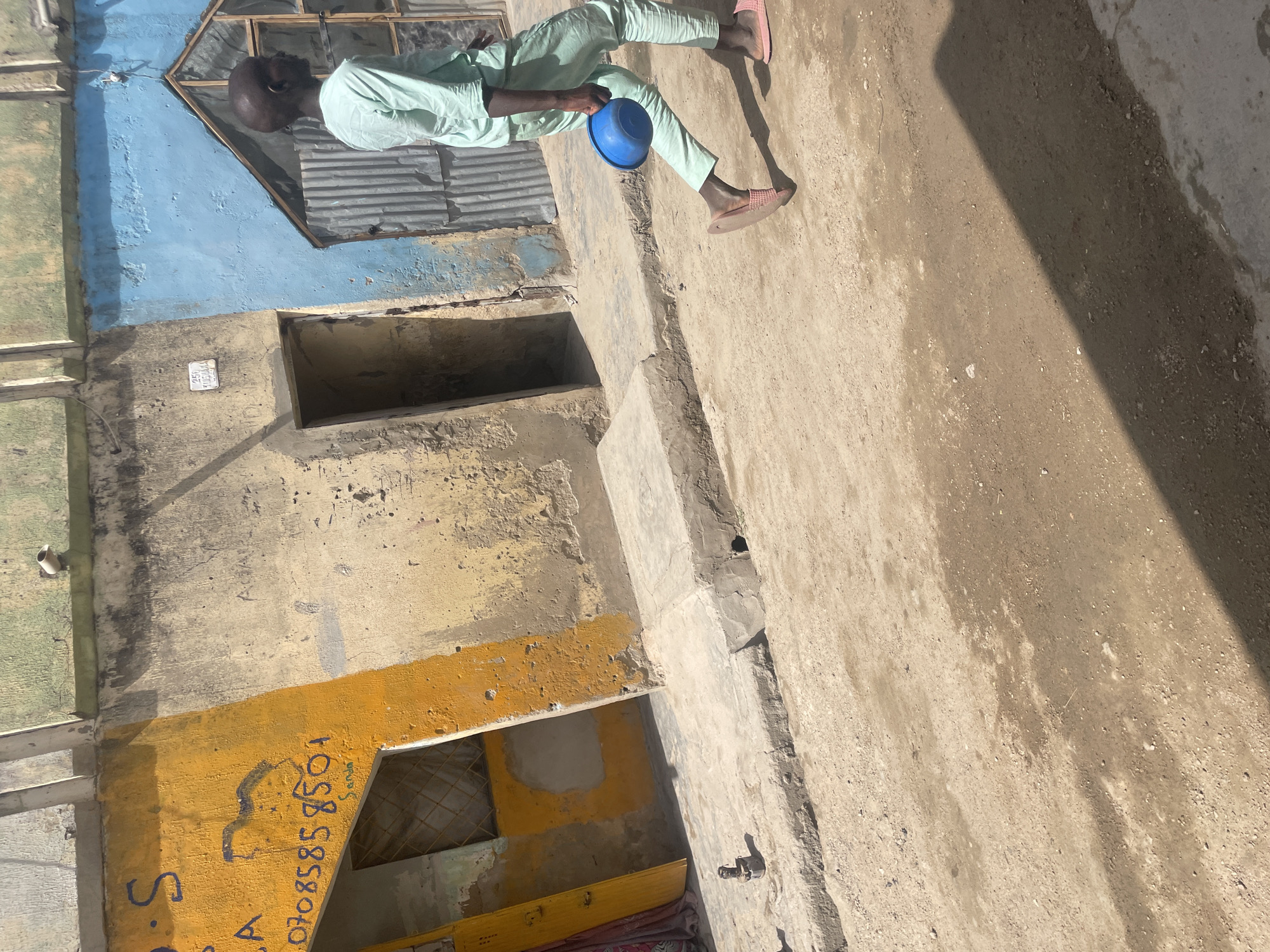 The image shows a person standing on a concrete surface next to a building in an urban environment. The person is holding a blue bowl. The building is painted in various colors, including blue, yellow, and green. The number 0708858501 is painted on the building. The location is Unknown, Unknown.