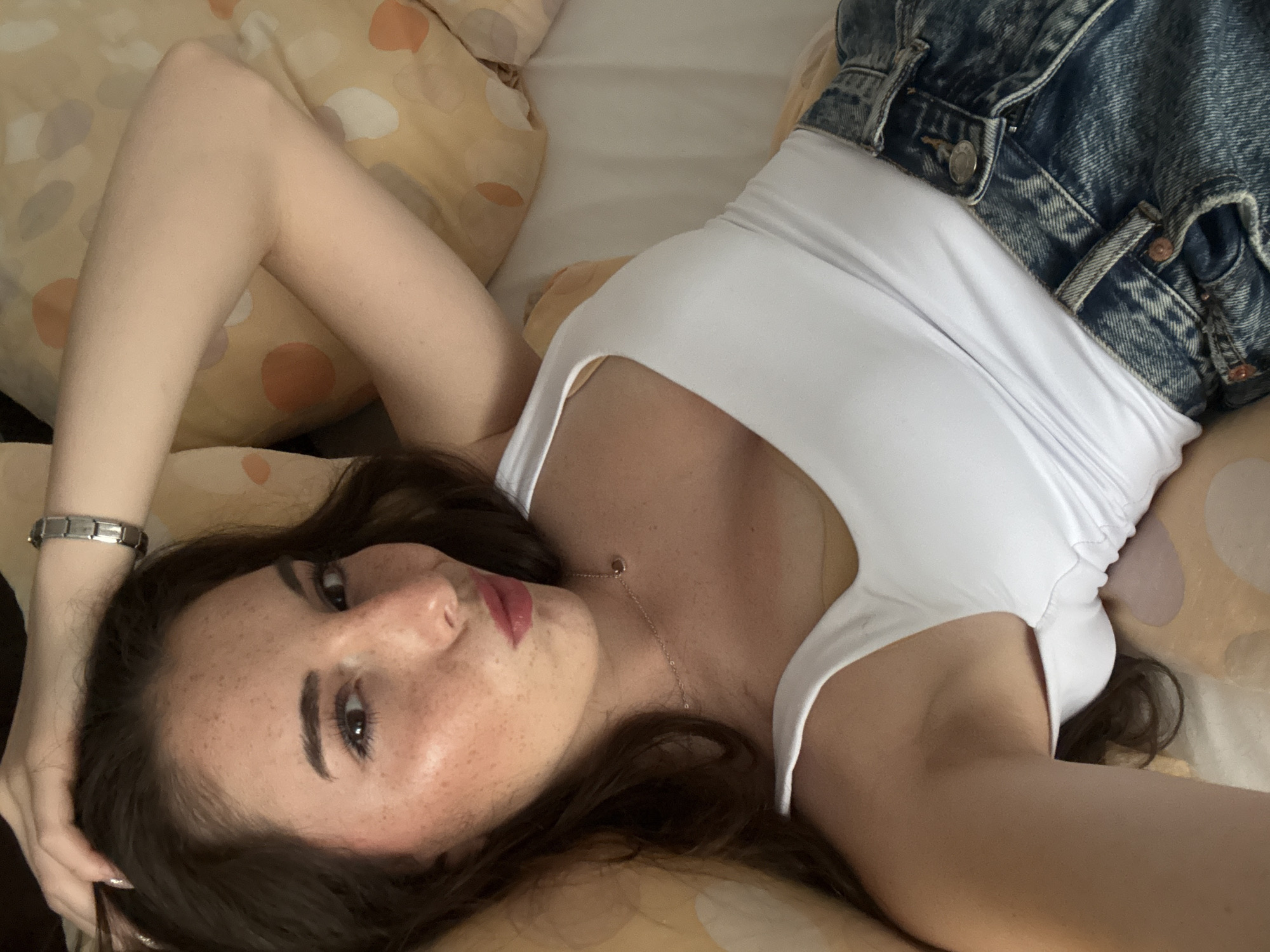 A young woman with long, dark hair and freckles lies on her back on a bed, resting her head on a pillow. She is wearing a white tank top and blue jeans. Her right arm is raised and bent, with her hand resting on her head. She is looking at the camera with her lips pursed. The background is a bed with pillows. There is no indication of the location being in Tazha, Bulgaria or any specific city.
