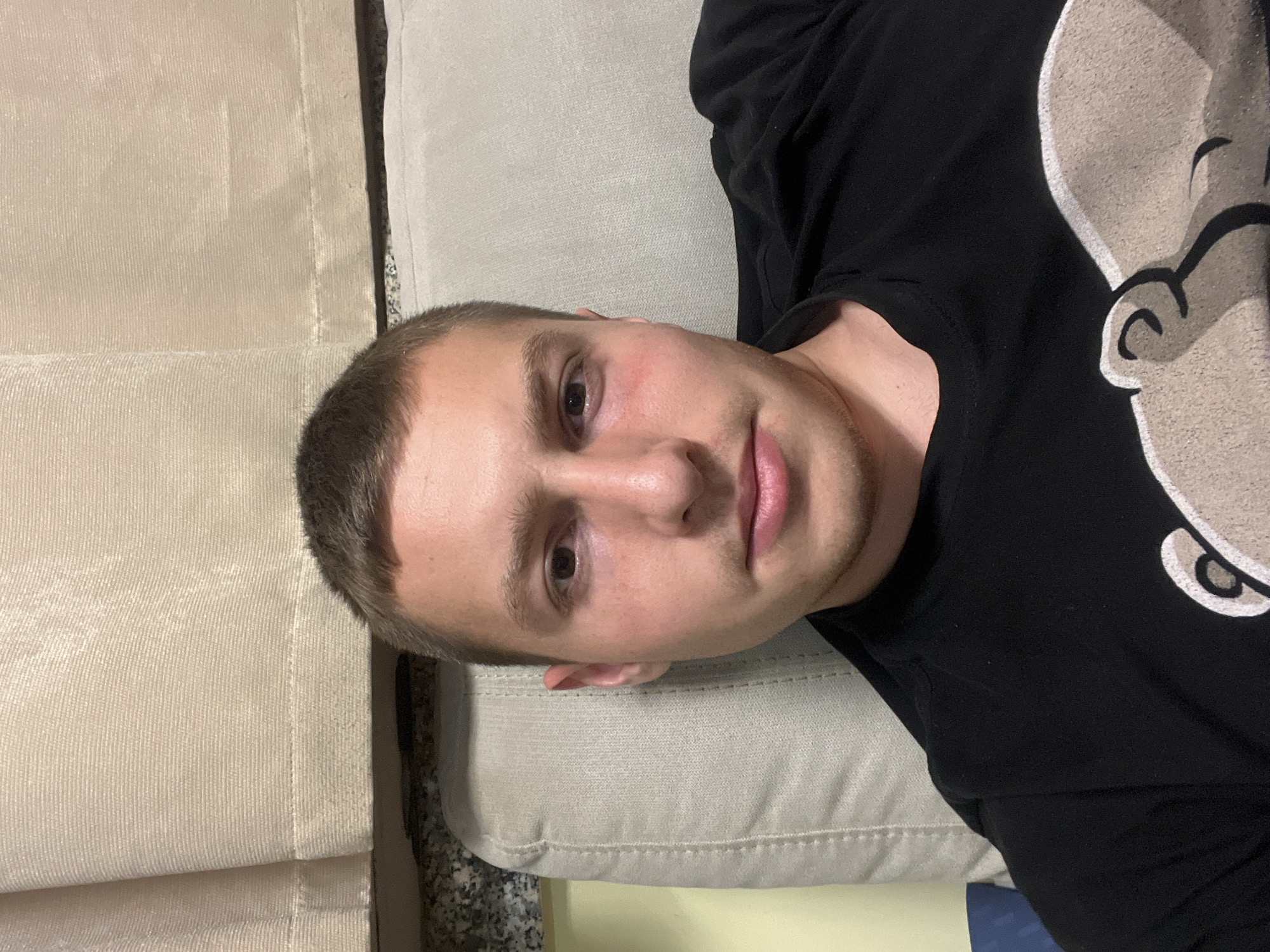 A light-skinned man with short brown hair and brown eyes lies on a cream-colored cushion or sofa. He is wearing a black shirt with a light-colored design featuring a cartoon character, possibly Popeye. The background includes beige fabric, suggesting curtains or drapes. There is no clear indication of the location in Burgas, Bulgaria, based on the provided image. There are no visible actions or interactions happening.