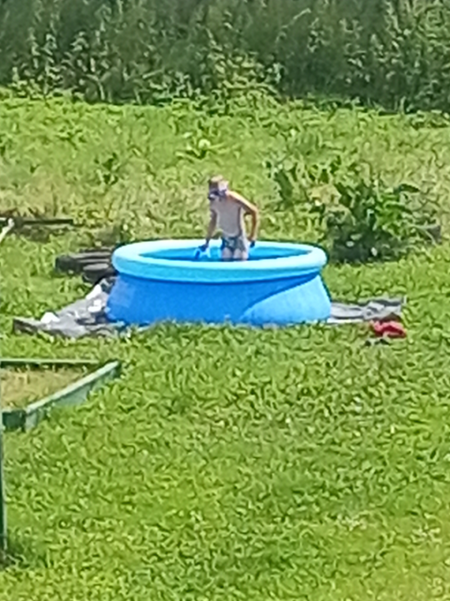 The image shows a young boy standing in a blue inflatable kiddie pool in a grassy area. The boy is wearing swimming trunks and appears to be wearing a snorkel mask. The pool is set up on a patch of ground that is partially covered by a gray tarp, and some other objects are visible around the pool. The background consists of green foliage and grass, suggesting an outdoor setting on a sunny day. The scene is likely from a backyard or park area in Shumilina, Belarus.