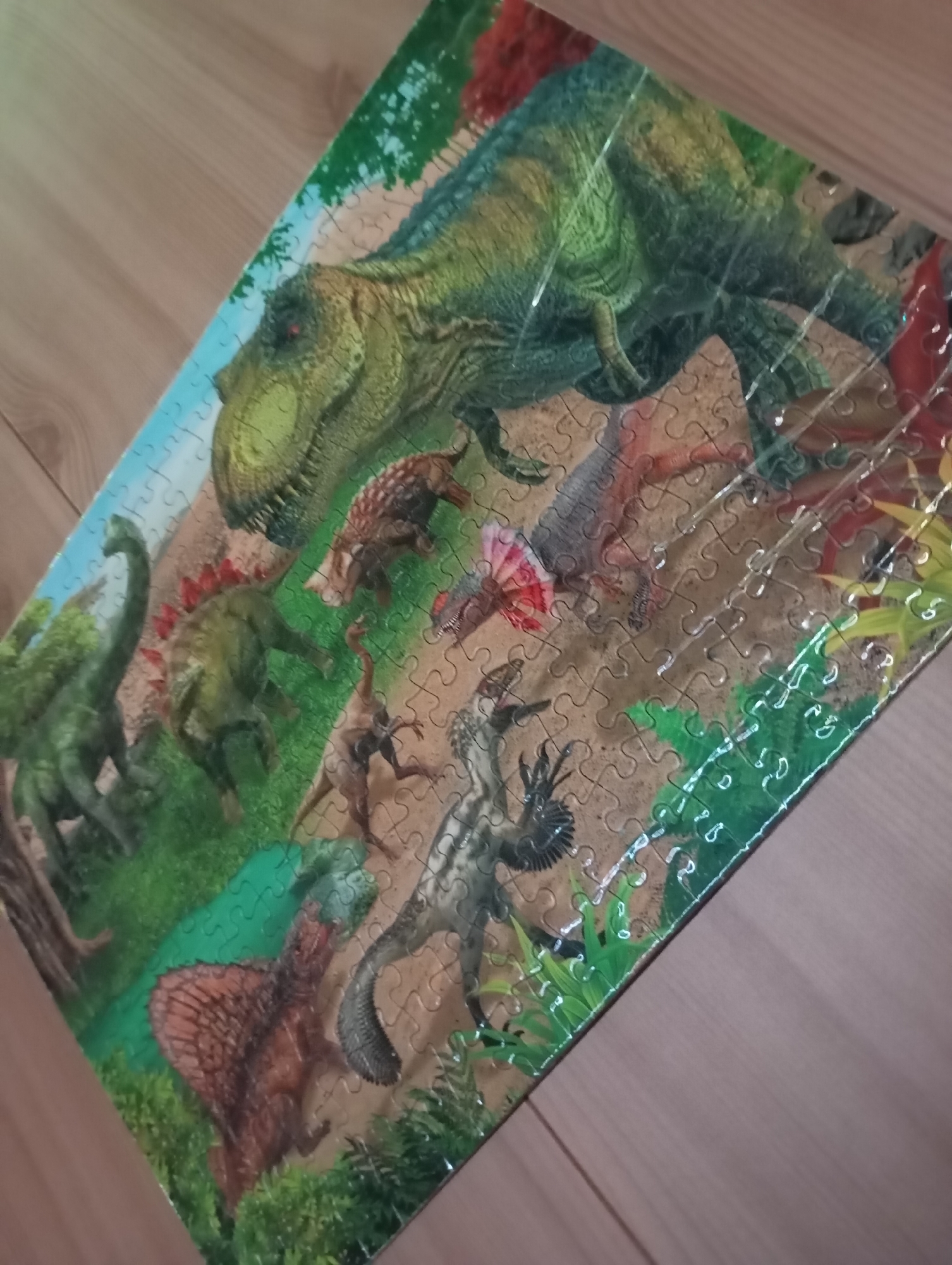 The image is a close-up shot of a completed jigsaw puzzle depicting various dinosaurs. The puzzle's primary subject is a large, green Tyrannosaurus Rex dominating the upper portion. Below the T-Rex are several other dinosaur species in a prehistoric landscape with green foliage, trees, and sandy-colored ground. The puzzle is on a wooden surface. The scene does not appear to be in any city.