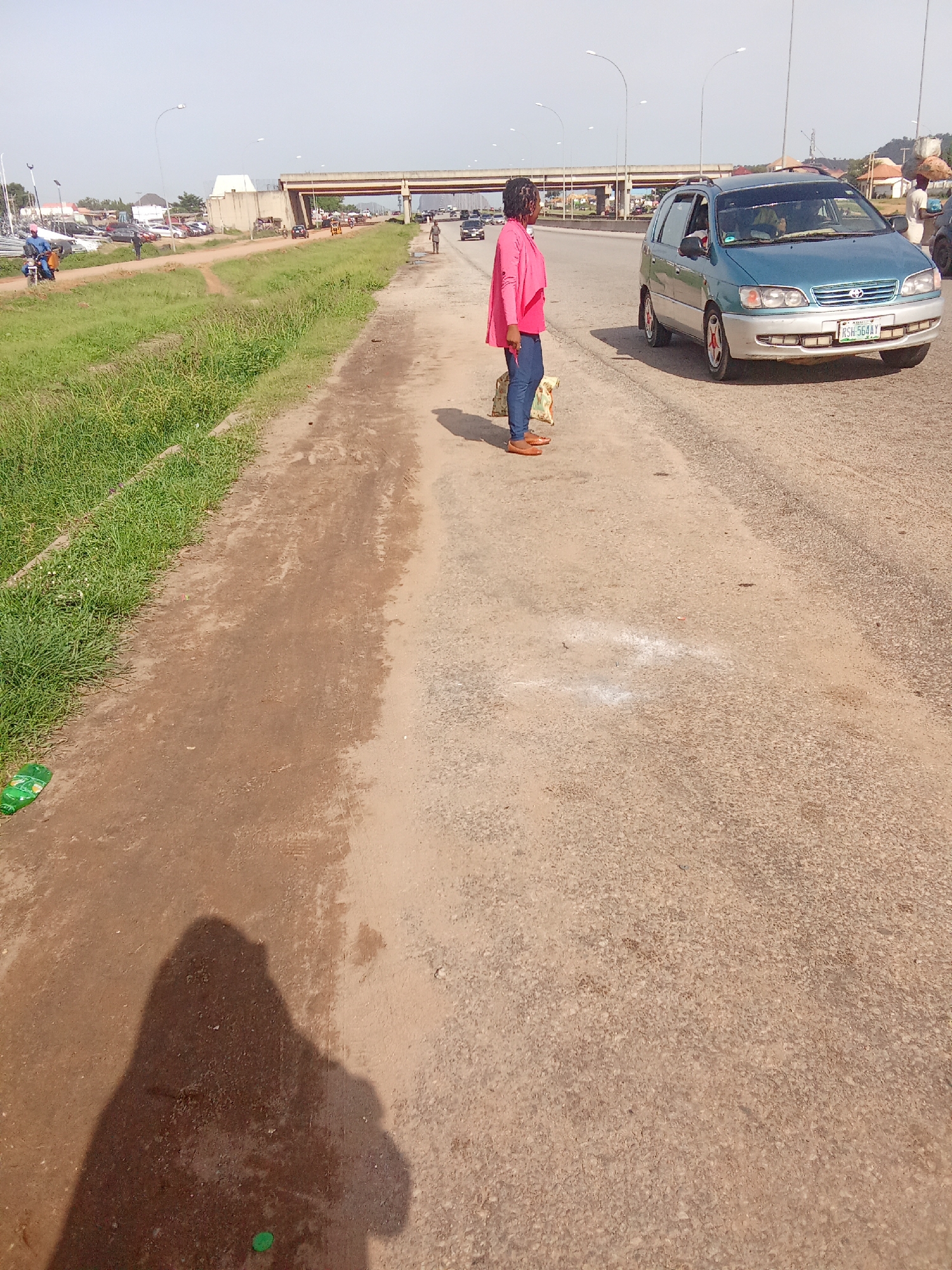 A person stands on a dusty roadside next to a teal minivan. The person is wearing a pink jacket and blue jeans and appears to be holding a bag. The minivan has a license plate that reads "RSH 564AY". In the background, there is an overpass and other vehicles on a highway. The scene appears to be a sunny day. The road is made of asphalt and dirt. There is also a grassy area to the left of the road. The overall setting suggests a roadside in Kagini, Nigeria.