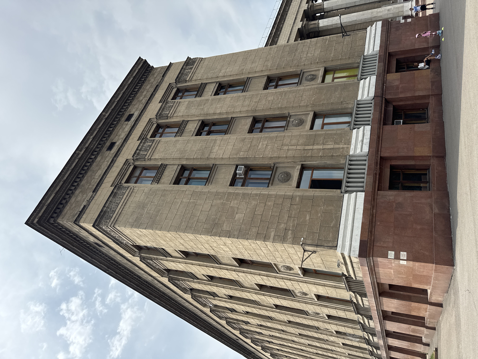 The image shows the exterior of the Almaty State Circus building in Almaty, Kazakhstan. The building is constructed of light-colored stone with a red granite base. It features multiple stories with numerous windows and decorative architectural elements. In the foreground, a paved area shows a few people. On the left, a person is sitting on the ledge of the building's base, wearing rollerblades. To the right of this person, two children are standing, one of whom is wearing a pink helmet and rollerblades, and the other is also wearing rollerblades. Further to the right, two adults are visible, one standing and the other on rollerblades. The sky above is partly cloudy.