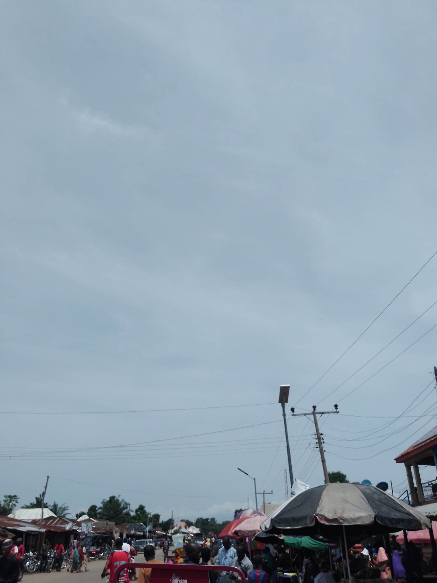 This is a street-level view of a bustling market scene in Serti, Nigeria, under an overcast sky. The foreground is dominated by a variety of market stalls, some covered by large, colorful umbrellas. People are engaged in various activities, including shopping, selling, and socializing. Motorcycles are parked along the roadside, indicating a common mode of transportation. Buildings with corrugated metal roofs line the street, suggesting a typical urban or semi-urban environment. Utility poles and power lines are visible, crisscrossing the sky. The atmosphere appears lively and active, characteristic of a marketplace.