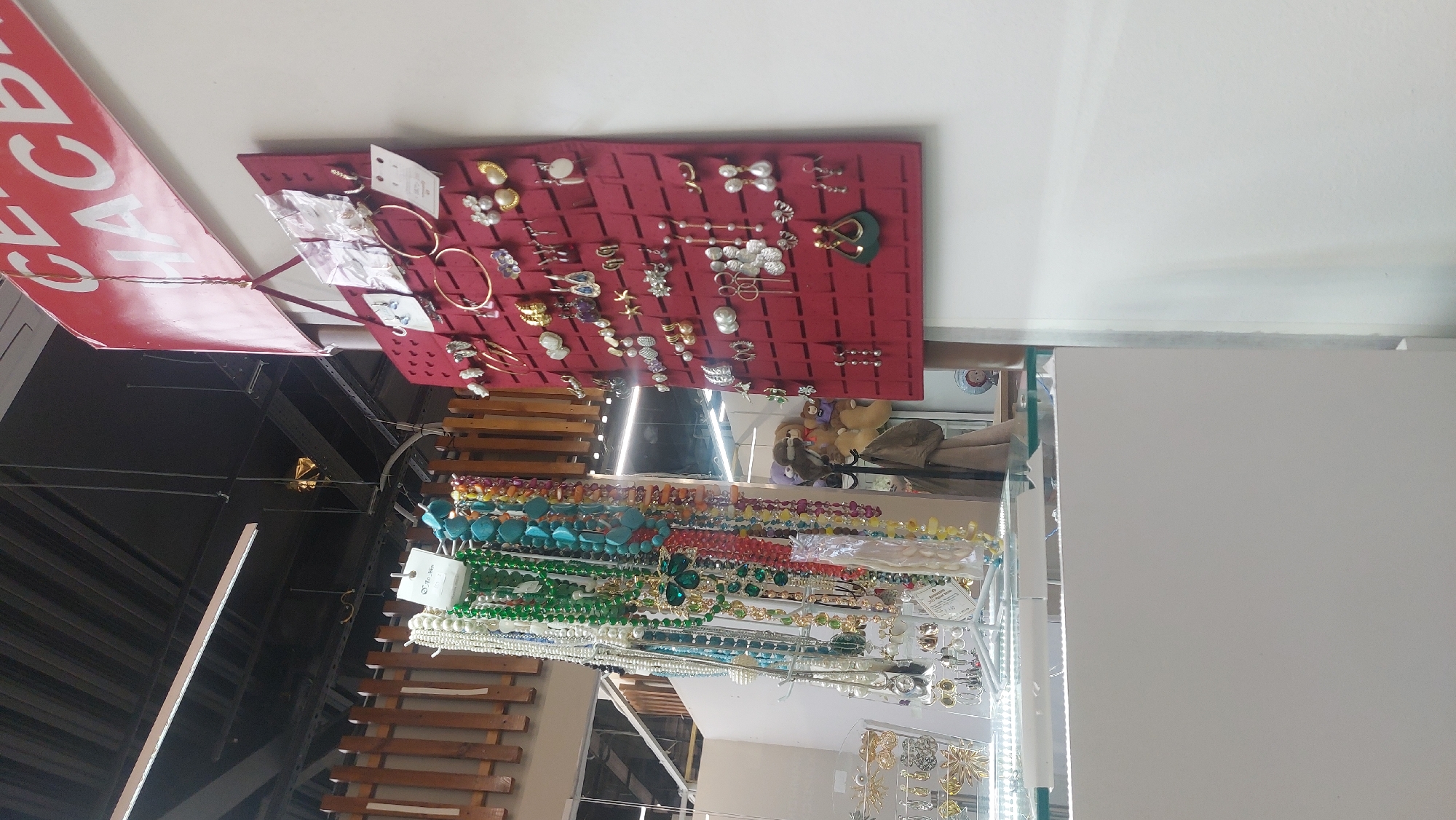 The image shows a store display in Novosibirsk, Russia. The display features a red fabric board with various earrings and rings attached. Below, there are multiple strands of beaded necklaces in various colors including turquoise, green, white and gold. The display is visible through a glass barrier. Background shows the interior of what appears to be a department store or market with scaffolding and ceiling structure visible. A red sale sign hangs above the jewelry.