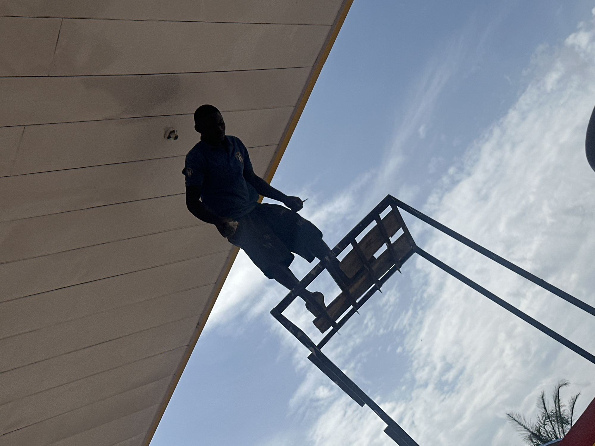 A low-angle, full-shot photograph taken in Barakin Lerawi, Nigeria, shows a dark-skinned man in silhouette standing on a metal scaffolding. The man, wearing a dark t-shirt and shorts, is leaning forward and appears to be holding a thin object, possibly a tool or a cigarette, in his right hand. He is positioned under a large, beige, corrugated overhang, likely a roof or canopy. The background is a clear blue sky with scattered white clouds. The scaffolding is constructed from dark metal beams and has wooden slats forming a platform. The image is taken from a low perspective, looking up at the man and the structures against the sky.