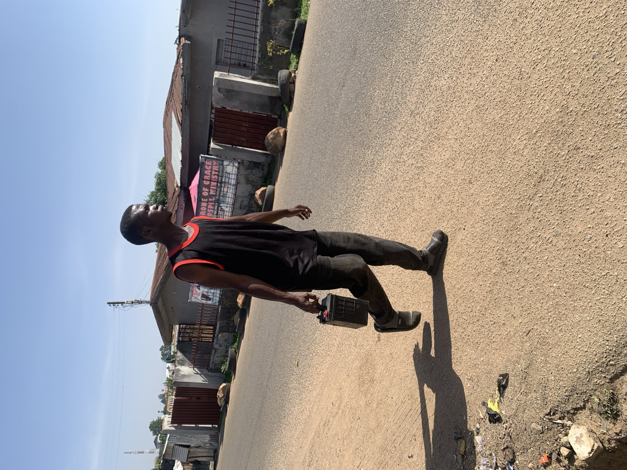 A man stands on a paved street in Jos, Nigeria, holding a car battery in his left hand. He is wearing a black sleeveless shirt with red trim, black pants, and dark gray shoes. The street has a grainy texture. To the left are buildings of varying construction, some with corrugated metal roofs. A sign for "ROME OF GRACE GOSPEL MINISTRY" is visible on one of the buildings. A utility pole stands in the distance. There is litter on the ground near the right edge of the street. The sky is clear and blue.