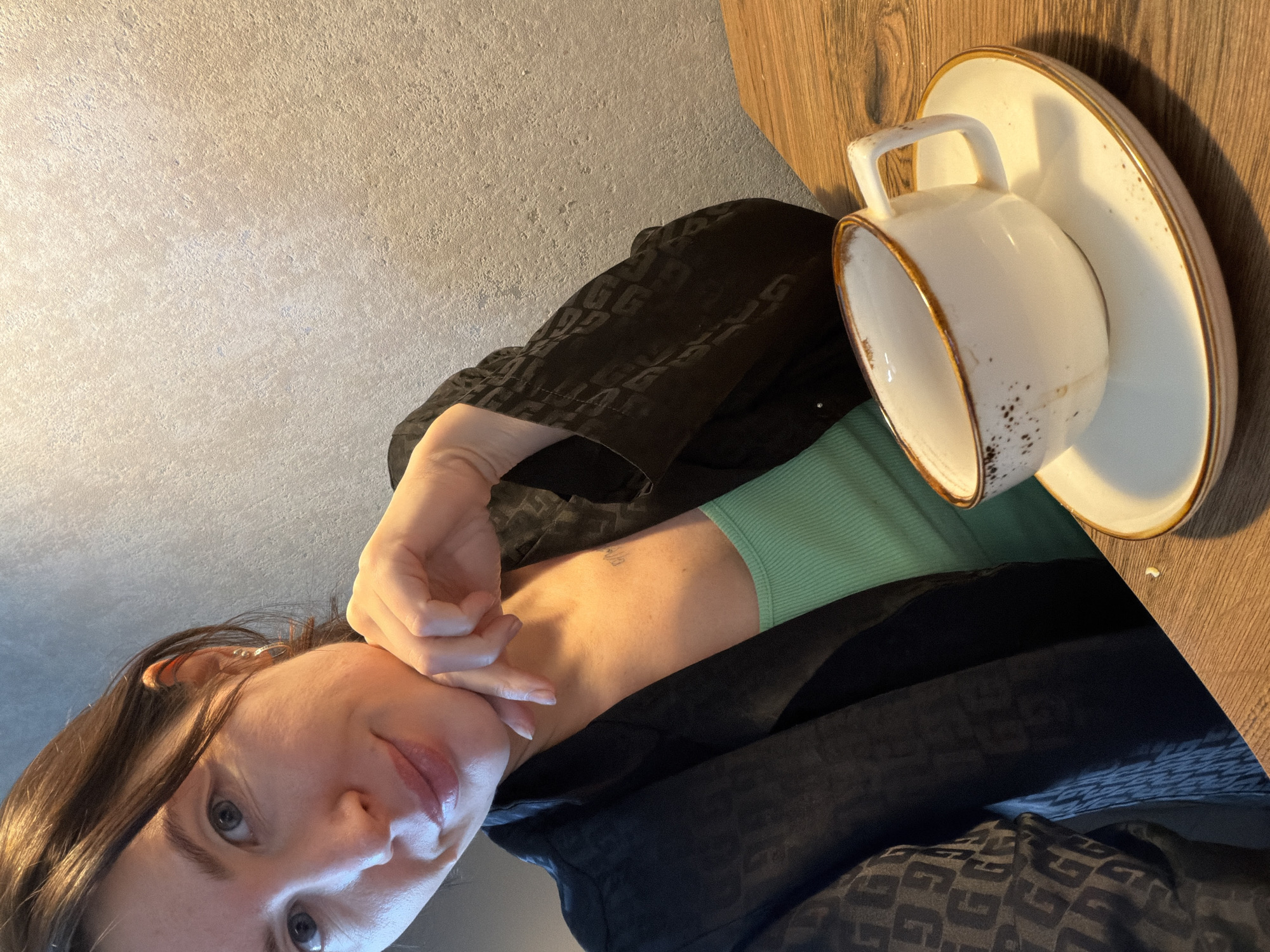 The image shows a woman seated at a wooden table, next to a white cup and saucer with a gold-colored rim. The woman has brown hair and blue eyes. She is wearing a green top and a patterned black jacket. She is holding her face with her left hand. The location is likely Perm, Russia, though this cannot be confirmed without additional context. The background consists of a textured wall.