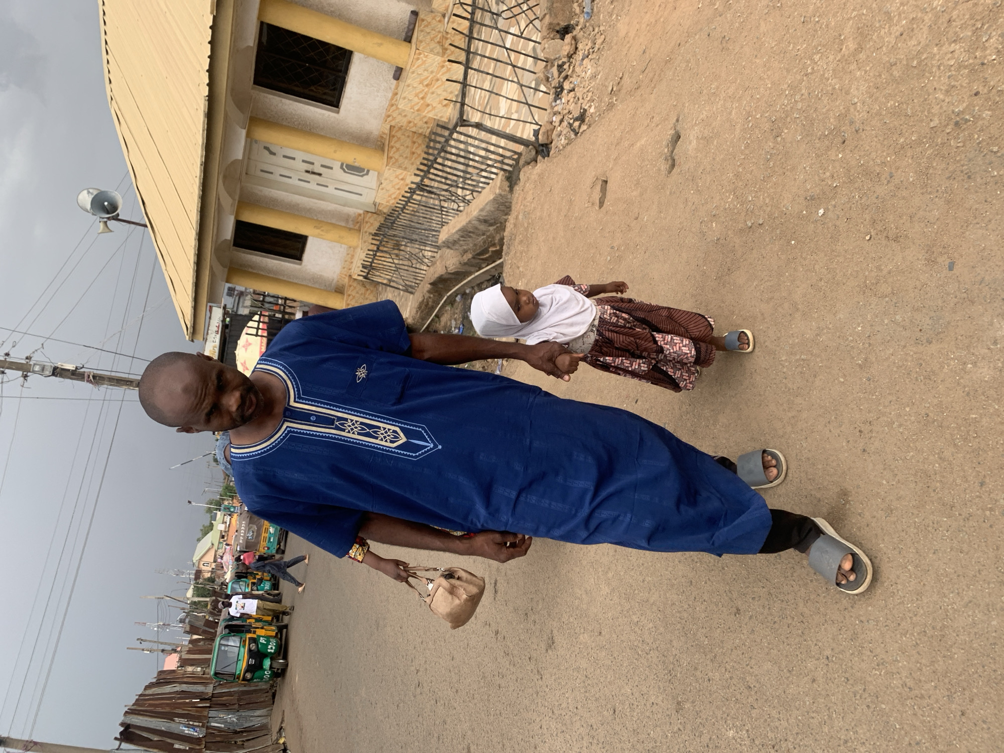 A man and a girl are walking on a street. The man is wearing a blue robe and sandals. The girl is wearing a white hijab and a brown patterned skirt. The man is holding the girl's hand and a purse. In the background, there is a building, vehicles, and power lines. Based on visual cues such as the style of clothing, types of vehicles (auto rickshaws/Tuk-tuks), and architecture, the location is likely in the city of Jos, Nigeria.