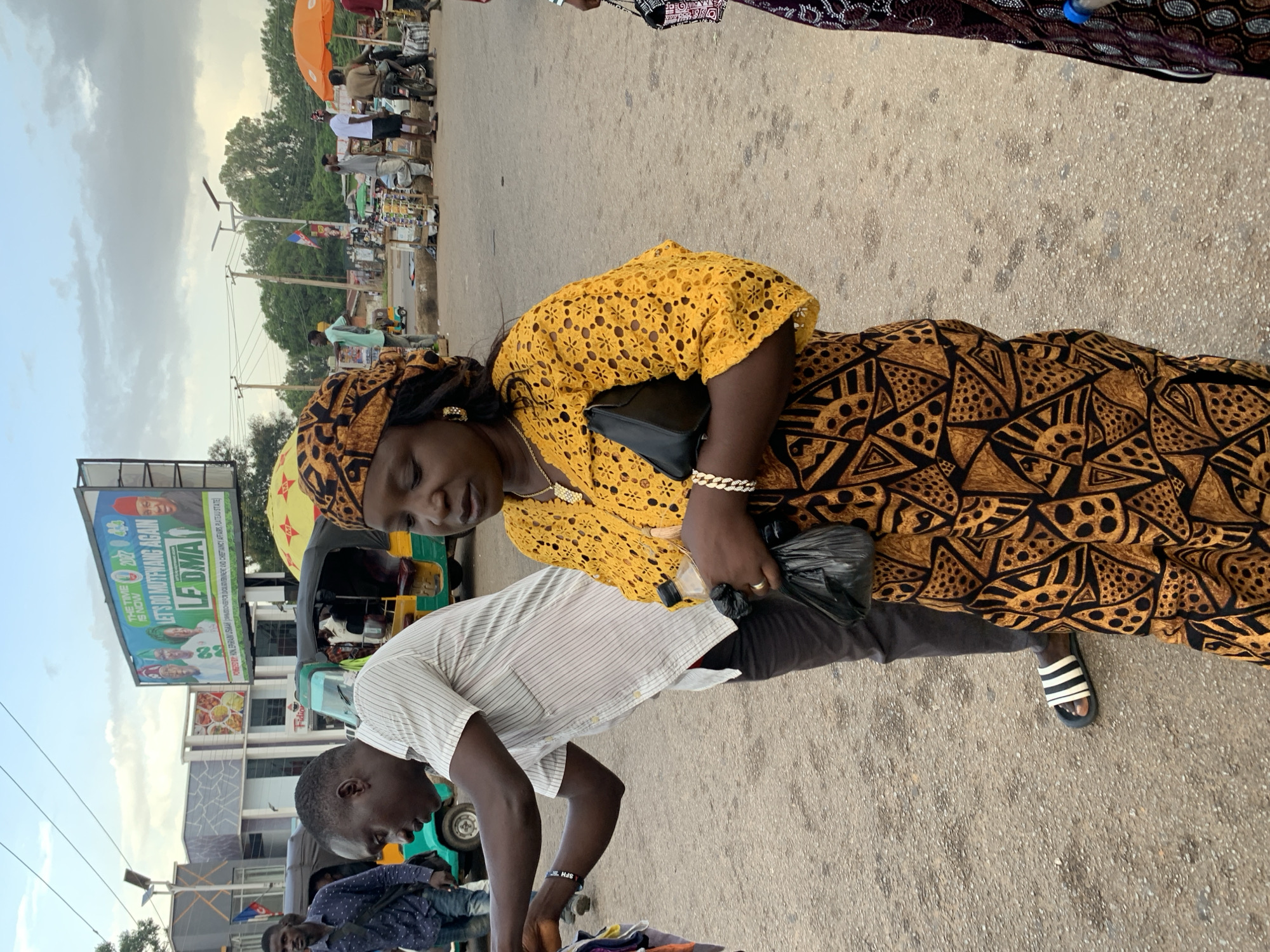 In Jos, Nigeria, on a paved road with scattered debris, a woman dressed in a yellow, intricately patterned dress and a matching headscarf is the central focus. She is holding a black clutch bag in her left hand and a plastic bottle in her right. A thick, gold-colored bracelet adorns her left wrist, and a gold necklace with a pendant hangs around her neck. Beside her, a man in a striped shirt and dark pants is engaged in a transaction, reaching towards something held by another person, partially visible on the left edge of the frame.
In the background, a large billboard displays a political advertisement with images of several individuals and text reading "THE TIME IS NOW" and "LET'S DO MUTFWANG AGAIN". Several three-wheeled vehicles, commonly known as Keke Napep, are parked or in motion. A yellow umbrella and an orange umbrella are also visible, suggesting a marketplace or roadside vendors. The sky is overcast with scattered clouds, and trees line the distant horizon. The overall scene depicts a busy urban environment with commercial activity.