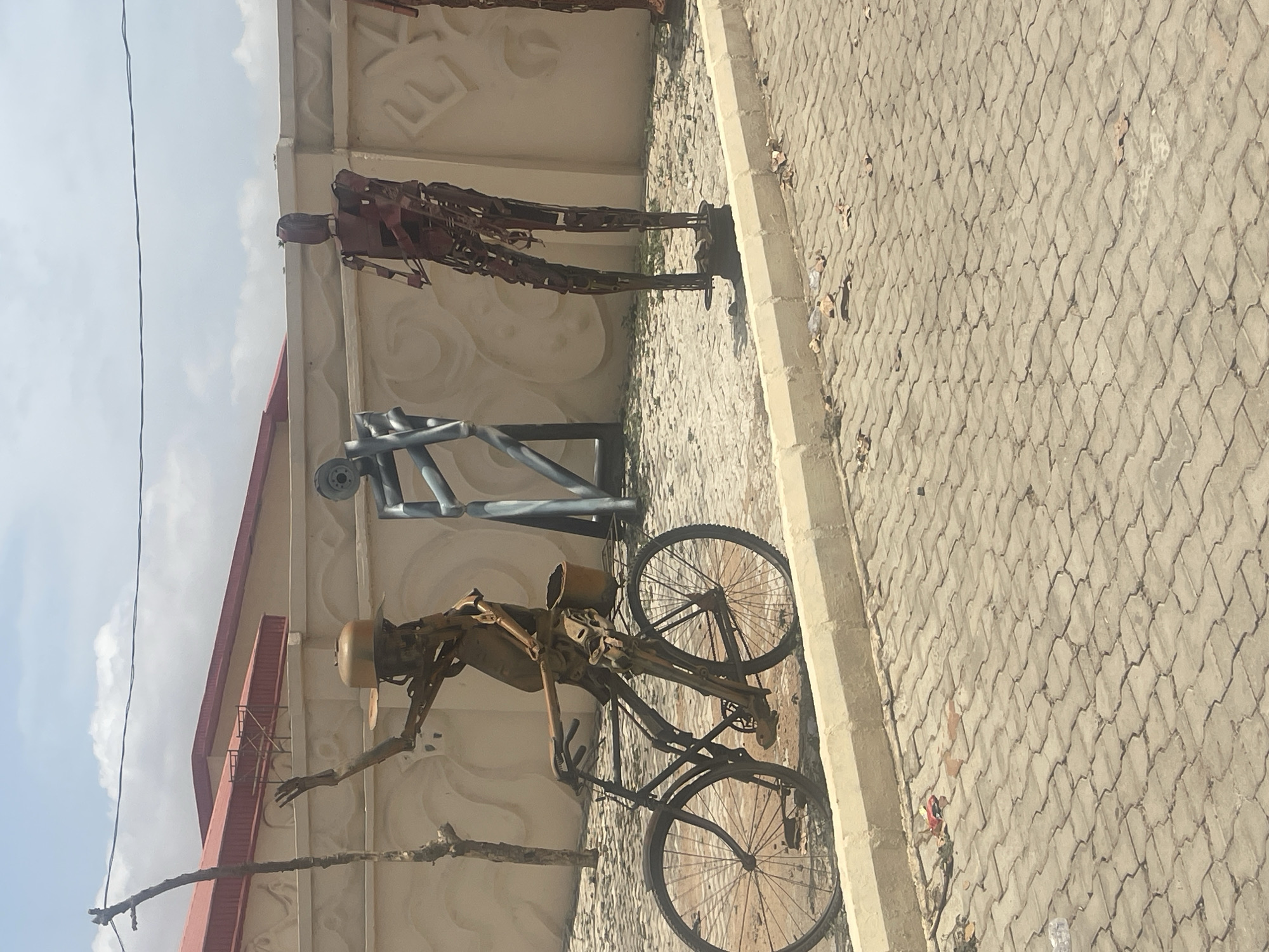 The image captures an outdoor scene featuring three distinct metal sculptures arranged on a paved ground in front of a building wall. The bright lighting and visible shadows indicate it is daytime under a clear to partly cloudy sky.
On the left, a detailed sculpture depicts a human-like figure riding a bicycle. This figure, crafted from repurposed metal components, is shown wearing a helmet and has a container attached to the rear of the bicycle. Extending from the left foreground, a thin, bare tree branch partially overlaps this sculpture.
Toward the center, an abstract sculpture stands, characterized by angular forms and a darker metallic finish. To its right, the third sculpture portrays a tall, slender human-like figure, also composed of various metal scraps, standing upright.
The ground beneath the sculptures is paved with light grey, interlocking concrete blocks, which show some scattered dry leaves and small debris. A light-colored concrete curb runs along the edge of this paved area, bordering the building. The building's wall, which serves as a backdrop, is light-toned and features subtle, decorative, embossed patterns. A reddish-brown roofline of the building is visible in the background. Overhead utility wires are seen stretching across the upper left portion of the sky.
The scene depicts a public art display or an outdoor exhibition space, consistent with the context of Abuja, Nigeria. There is no visible text in the image.