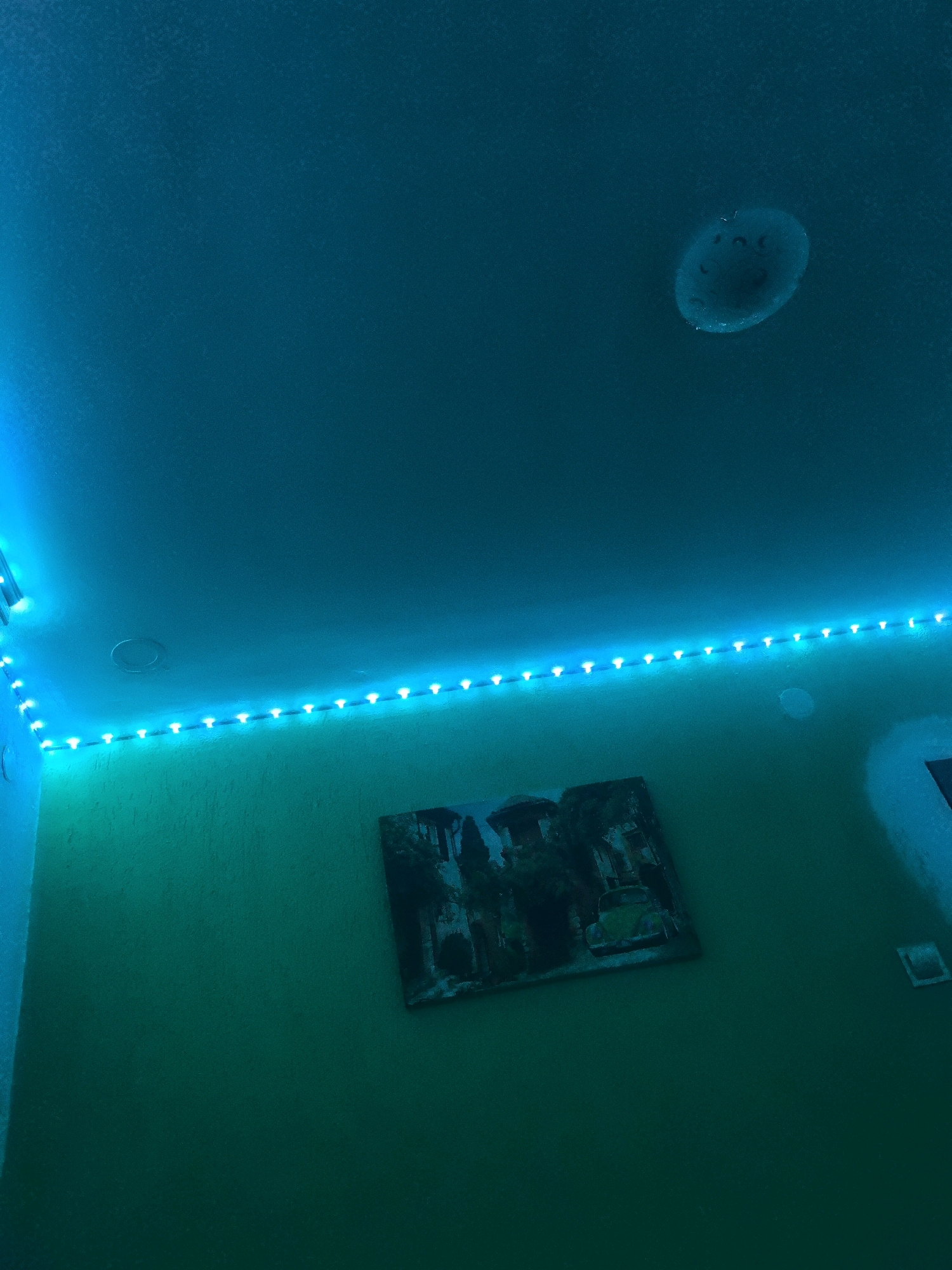 The image shows a corner of a room with walls and ceiling illuminated by blue and green LED strip lights. There is a framed picture of a landscape with ancient ruins and a yellow car hanging on the green wall. A circular light fixture is visible on the blue-lit ceiling. The overall impression is of a modern, moodily lit interior. The provided information about the city of Kotel, Bulgaria, is extraneous to the visual content of the image.
