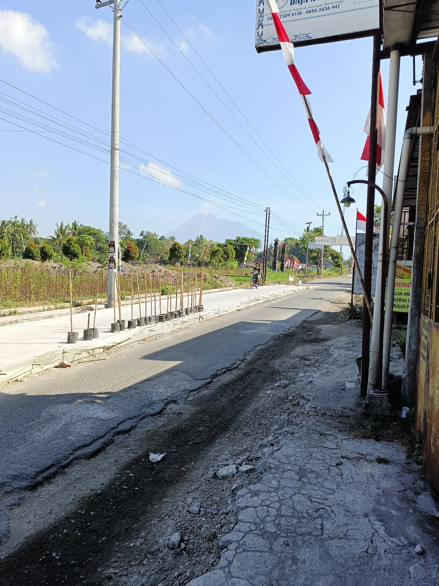 The image depicts a roadside scene in Sukorini, Indonesia, under a clear blue sky with scattered clouds. A paved road, showing signs of wear and cracking, runs from the foreground into the distance. To the left of the road, a sidewalk area is lined with numerous young trees or saplings in black plastic bags, each supported by a wooden stake. A tall utility pole with numerous wires stands prominently on the left side of the image. Further down the road, a distant volcano is visible, its peak shrouded in mist.
On the right side of the road, a building with a yellow, textured wall stands. A sign is attached to the building, displaying red and white Indonesian flags. Another Indonesian flag, a long banner of alternating red and white stripes, hangs from a pole in front of the building. Various posts and pipes are also present near the building. In the middle ground, a person rides a motorcycle on the road, heading away from the camera. The surrounding environment includes green vegetation, trees, and an undeveloped plot of land with fencing. The overall impression is of a rural or semi-urban environment on a sunny day, with elements of local culture and infrastructure.