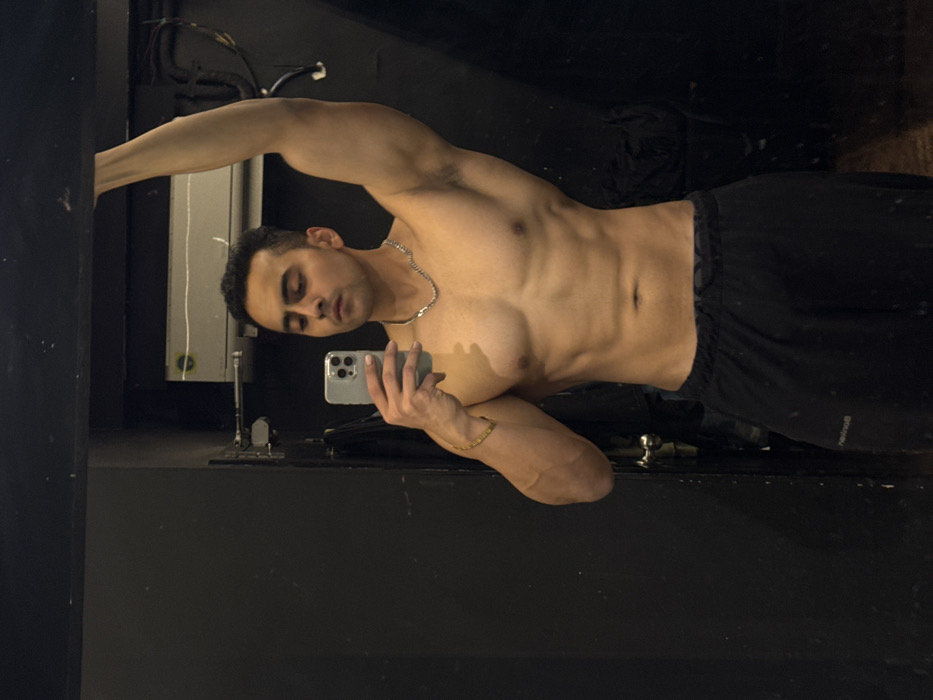 A shirtless man with tan skin and a silver necklace stands angled in front of a mirror, using his phone to take a picture of himself. He wears dark workout shorts, and his physique shows toned abdominal muscles and well-defined arms. His face is angled slightly downward, and his eyes are closed. The background is dimly lit, with a black wall and a reflective surface creating the mirror effect. There are some wires, perhaps from a lighting fixture, hanging from the ceiling in the background.