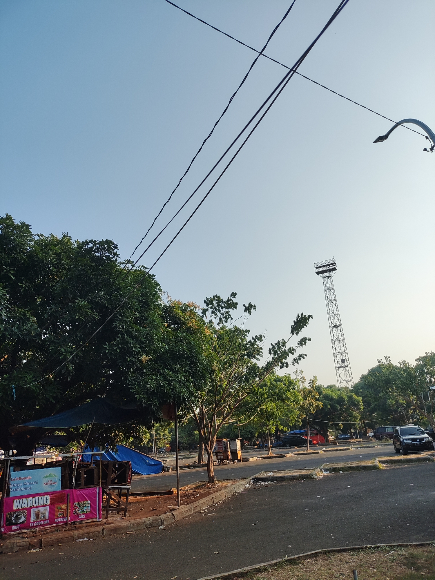 This is a photograph of an outdoor scene, likely a parking lot or open area adjacent to a road. The time of day appears to be late afternoon or early evening, given the lighting conditions. A tall, lattice-like tower, possibly a cell tower or floodlight tower, is visible in the background, reaching towards the clear blue sky. Power lines crisscross the upper portion of the frame, and a streetlamp is mounted on a curved pole on the right.
In the foreground and mid-ground, several elements suggest a temporary or informal commercial area. There are several stalls or small shops, some covered by blue tarps. One stall prominently displays a pink banner with the word "WARUNG" (Indonesian for shop or stall) and images of coffee and other beverages, indicating it sells food and drinks. Signs with text in Indonesian, such as "Le Minerale," are also visible.
A paved road or parking lot dominates the lower half of the image, with several parked vehicles, including a black SUV. Green trees are abundant throughout the scene, providing shade and natural elements. The ground in the parking area appears to be asphalt, with some patches of grass and debris. The overall environment seems to be a semi-urban or suburban area in Cirebon, Indonesia, characterized by a blend of infrastructure, commerce, and natural vegetation. There are no people clearly visible in the image.