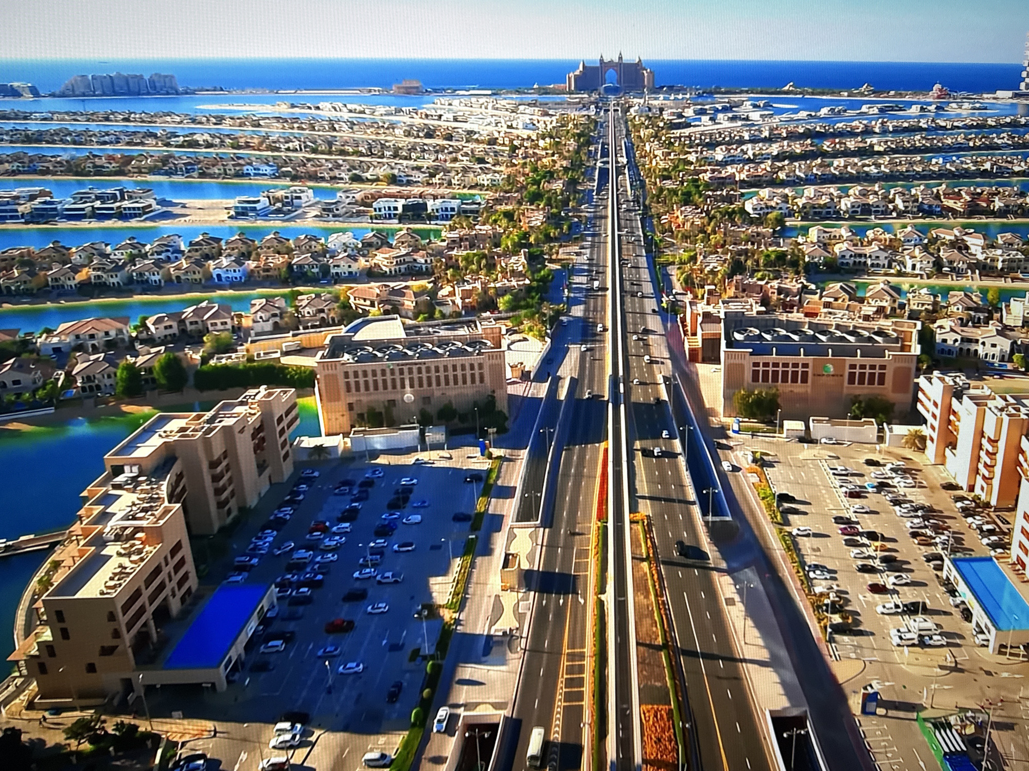 This is an aerial drone shot of a large-scale infrastructure project in a coastal city. The scene features a multi-lane highway with several vehicles driving on it, flanked by residential buildings, canals, and green spaces. In the distance, a massive hotel complex is visible on a man-made island, with the ocean stretching out beyond. The architecture is modern and luxurious, suggesting a high-end resort or residential area. The location is Dubai, United Arab Emirates, not Burgas, Bulgaria. There are no people visible in the image.