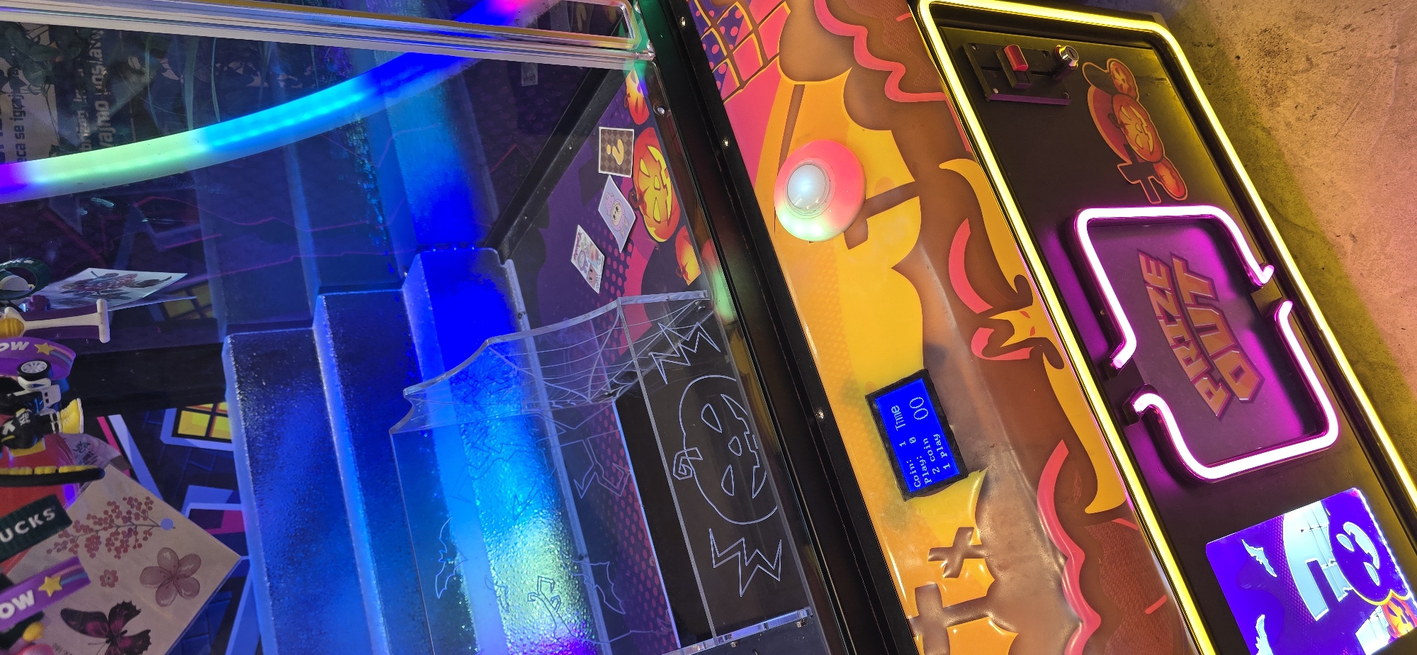 The image shows a close-up view of a "Prize Out" arcade game. The game has a Halloween theme with images of pumpkins and spiders. There are prizes inside the game, and an LCD screen that has the pricing information. The environment includes a neon light.