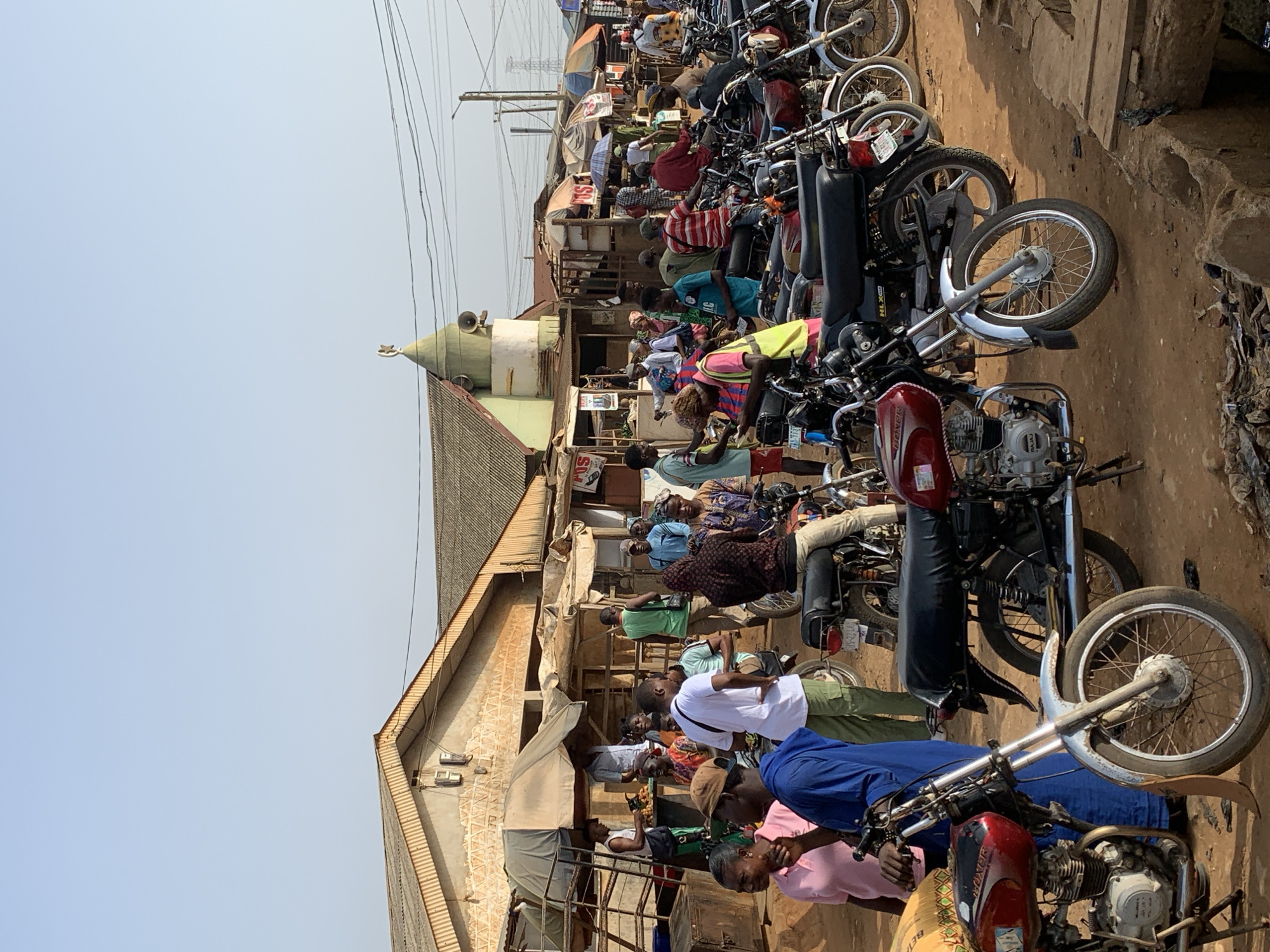 The image depicts a street scene in Ikorodu, Nigeria. A row of parked motorcycles lines the side of a street, with people seated on some of them. Adjacent to the motorcycles are buildings, including one with a green-domed structure, likely a mosque. People are gathered in front of the buildings. The weather appears sunny, and electrical wires are visible overhead.