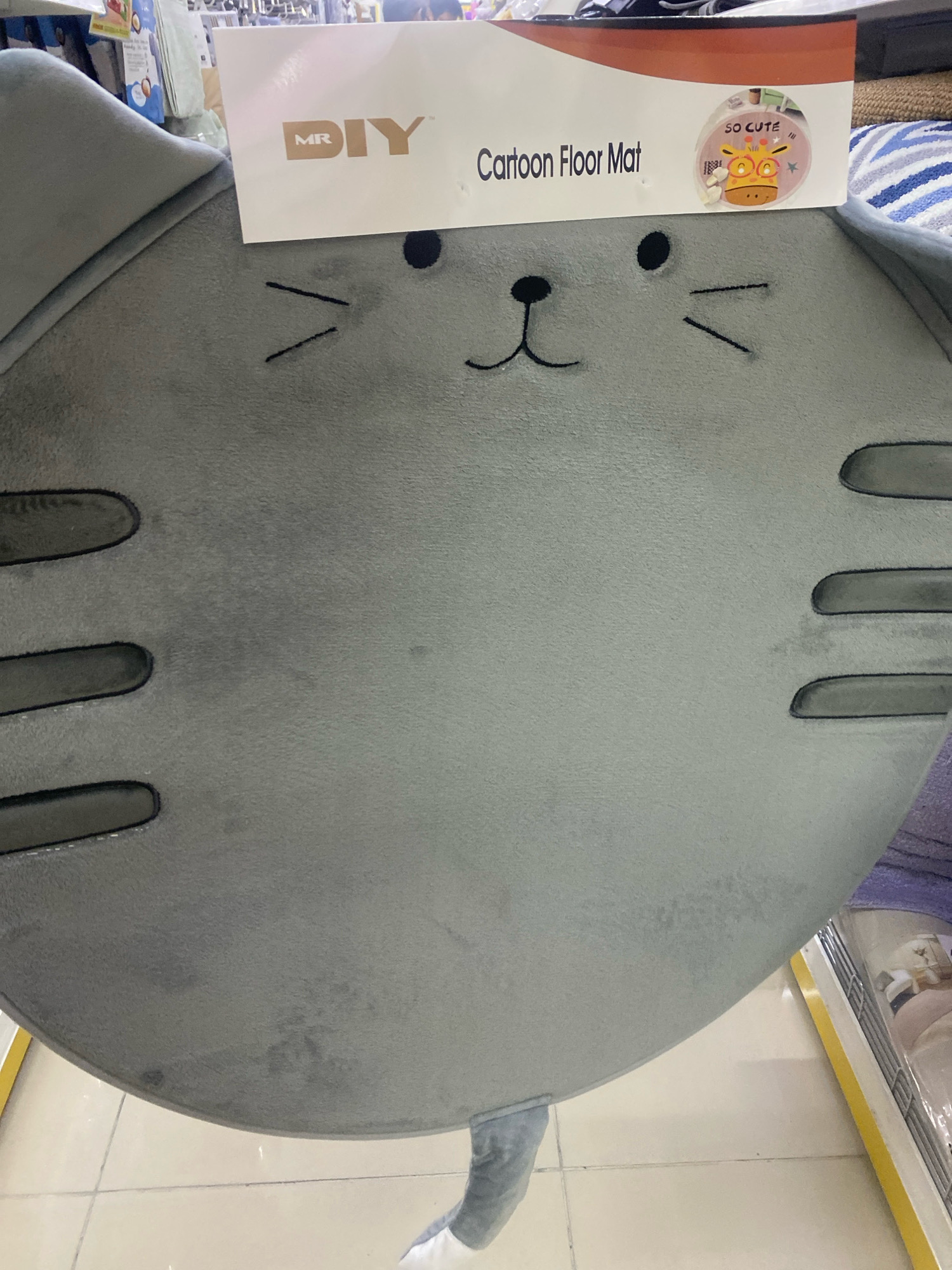 A grey, plush floor mat shaped like a cartoon cat's face is displayed on a retail shelf in what appears to be a MR DIY store in Kuala Lumpur, Malaysia. The mat features embroidered eyes, nose, and whiskers, and has dark grey oval shapes on its sides that resemble paws. A white cardboard tag with the MR DIY logo and "Cartoon Floor Mat" printed on it is attached to the top of the mat. A small circular sticker on the tag shows a cartoon giraffe wearing glasses and the words "SO CUTE". The background of the image shows other retail items and shelving. The mat is the primary subject and is positioned centrally in the frame, with its lower portion extending out of view.