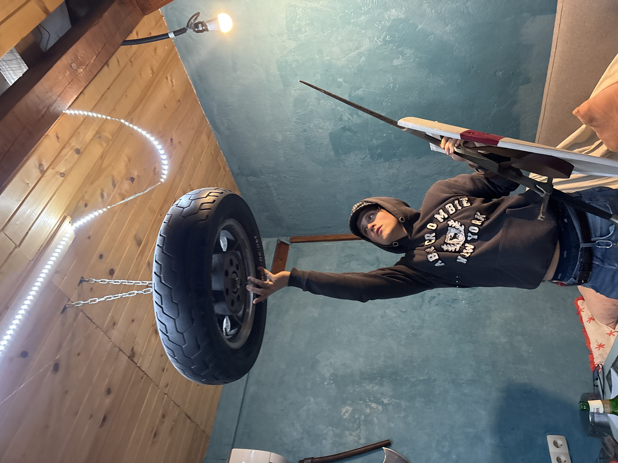 A low-angle shot of a male person in a black hoodie, jeans, and a dark hat, standing indoors. The person is holding an ironing board in their right hand and touching a car tire suspended from the ceiling with their left hand. The tire is attached to the ceiling by metal chains. The ceiling is painted blue, and the walls are wooden. A strip of LED lights is attached to the wooden walls. An axe is leaning against the wall. There is no indication of the city of Verinsko, Bulgaria in the image.