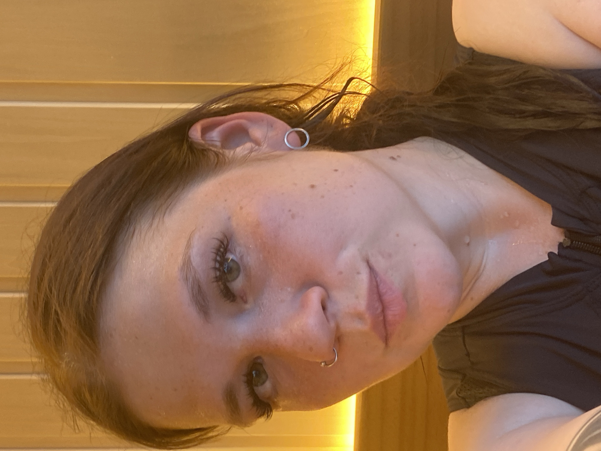 A close-up, head-and-shoulders portrait of a young Caucasian woman with brown hair and green eyes is presented. She is looking directly at the camera with a slight pout. Her face is adorned with freckles and she is wearing false eyelashes. She has a silver hoop nose ring and a silver hoop earring in her right ear. The background consists of vertical wooden panels, illuminated by warm lighting from above, suggesting an indoor setting possibly in Votkinsk, Russia. The woman is wearing a dark, sleeveless top with a zipper, and her skin appears slightly dewy, possibly from sweat.